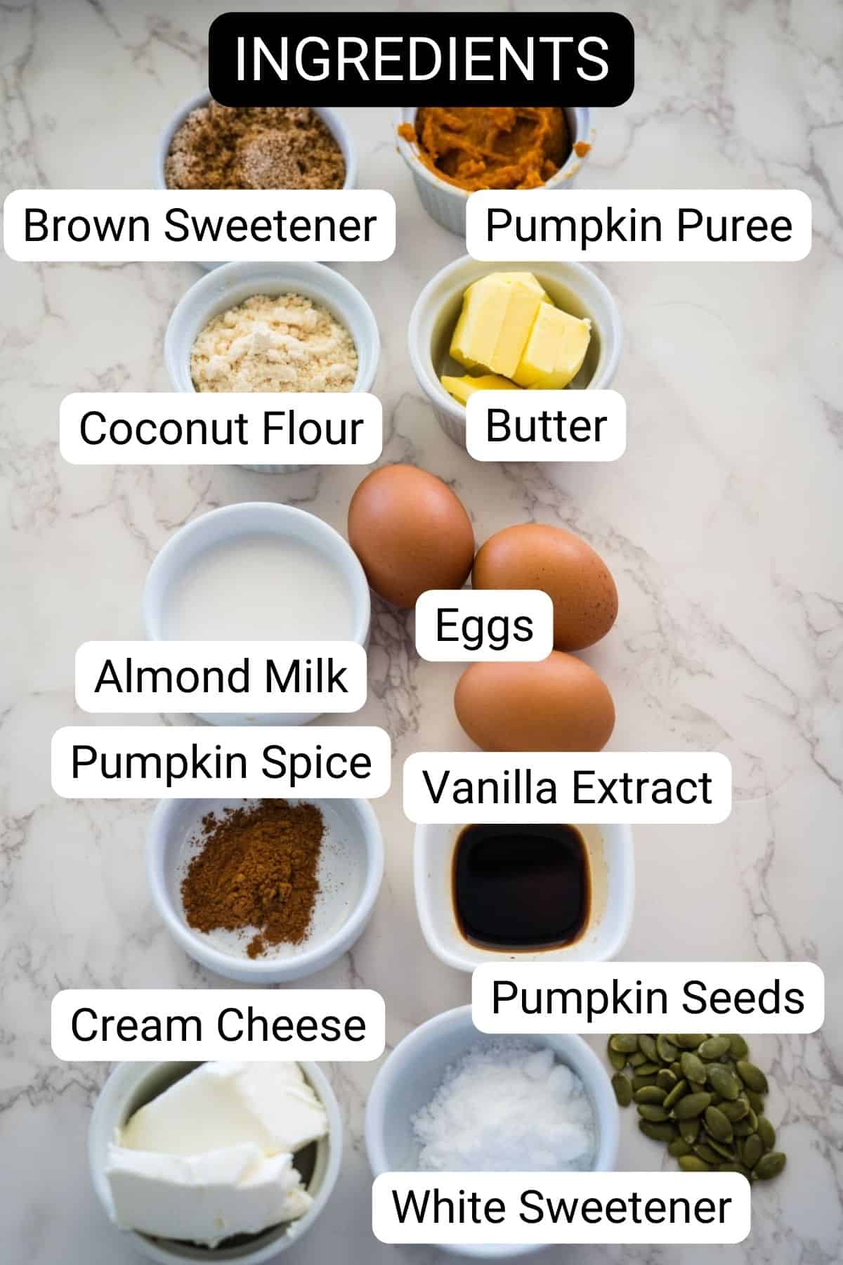 Variety of labeled ingredients in bowls on a marble surface, including brown sweetener, pumpkin puree, coconut flour, butter, eggs, almond milk, pumpkin spice, vanilla extract, cream cheese, pumpkin seeds, and white sweetener.