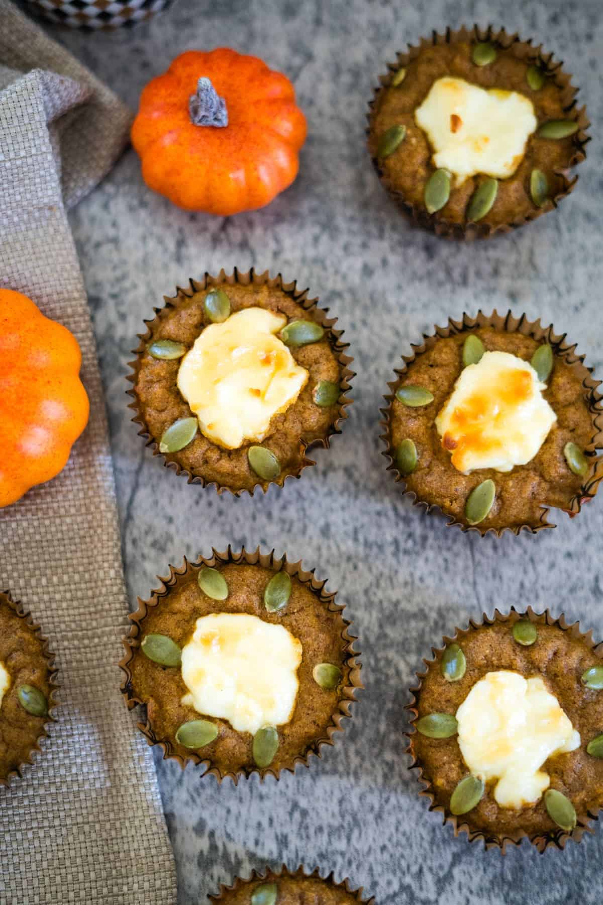 Pumpkin muffins topped with cream cheese and pumpkin seeds are arranged on a gray surface with small orange pumpkins and a textured cloth nearby.