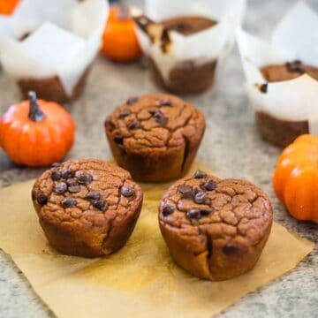 Pumpkin chocolate chip muffins on parchment paper, surrounded by small decorative pumpkins and additional muffins in the background.