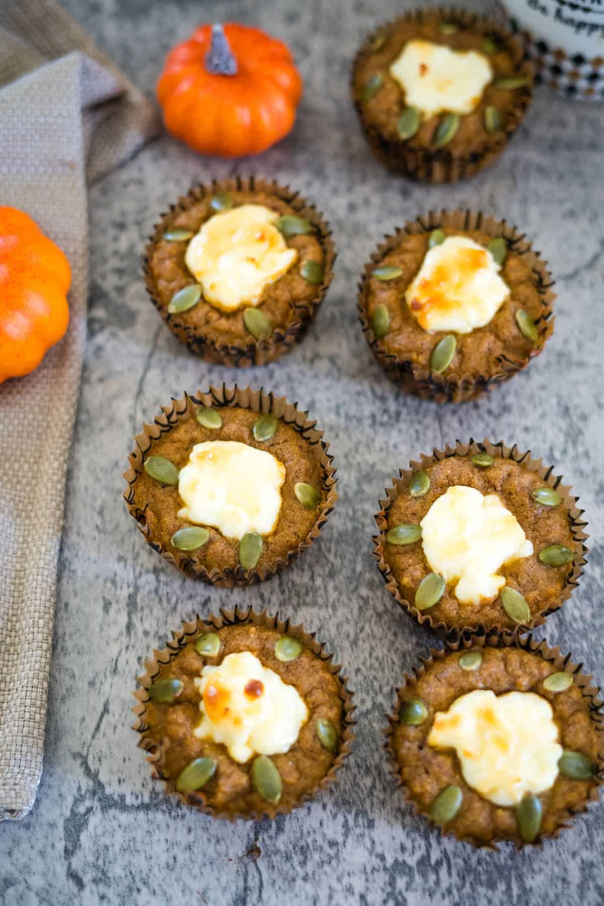 Six keto pumpkin cheesecake muffins topped with pumpkin seeds and dollops of cream cheese are arranged on a gray surface. Small decorative pumpkins and a beige cloth are nearby.