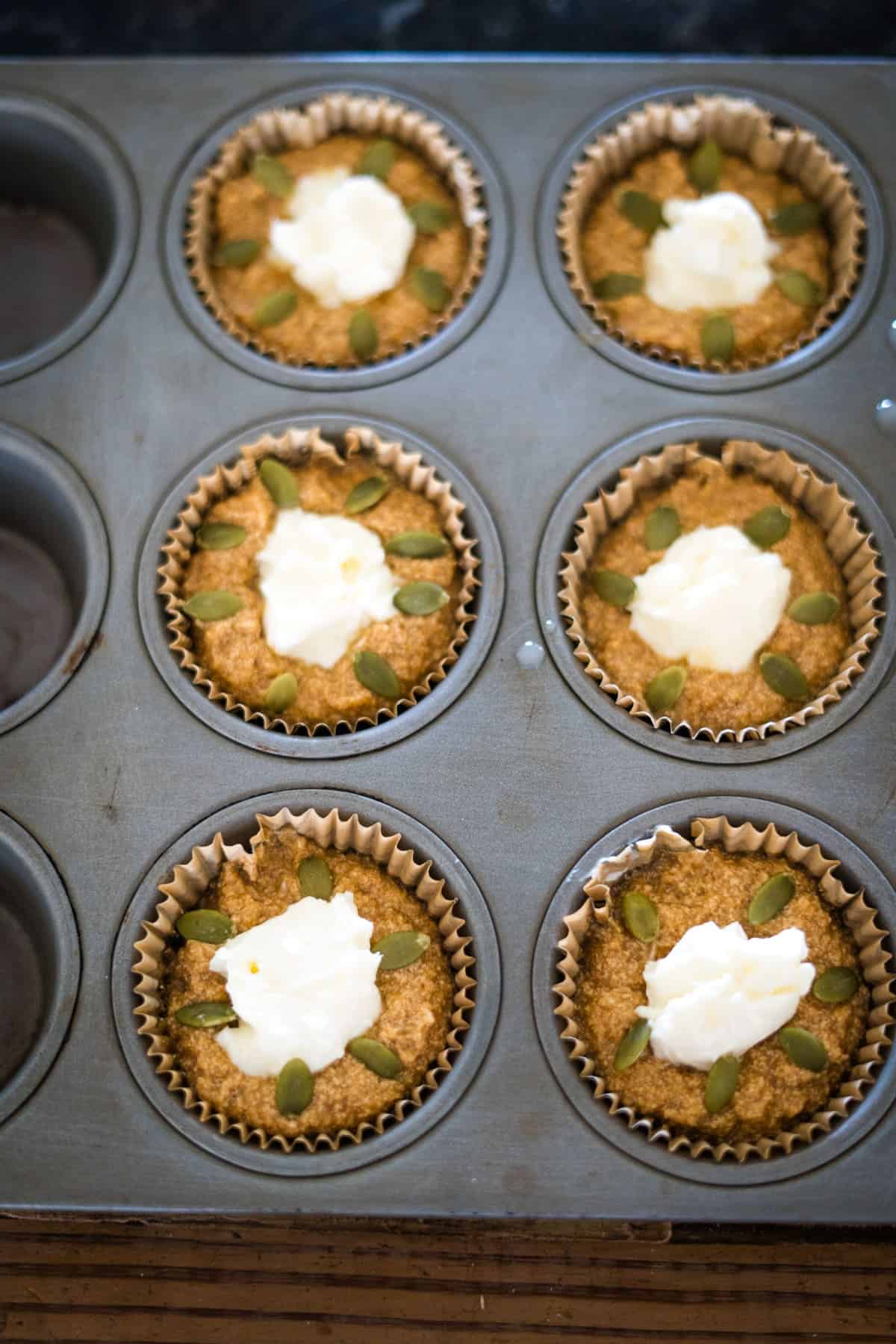 A muffin tray with six keto pumpkin cheesecake muffins topped with cream cheese and pumpkin seeds, placed in paper liners.