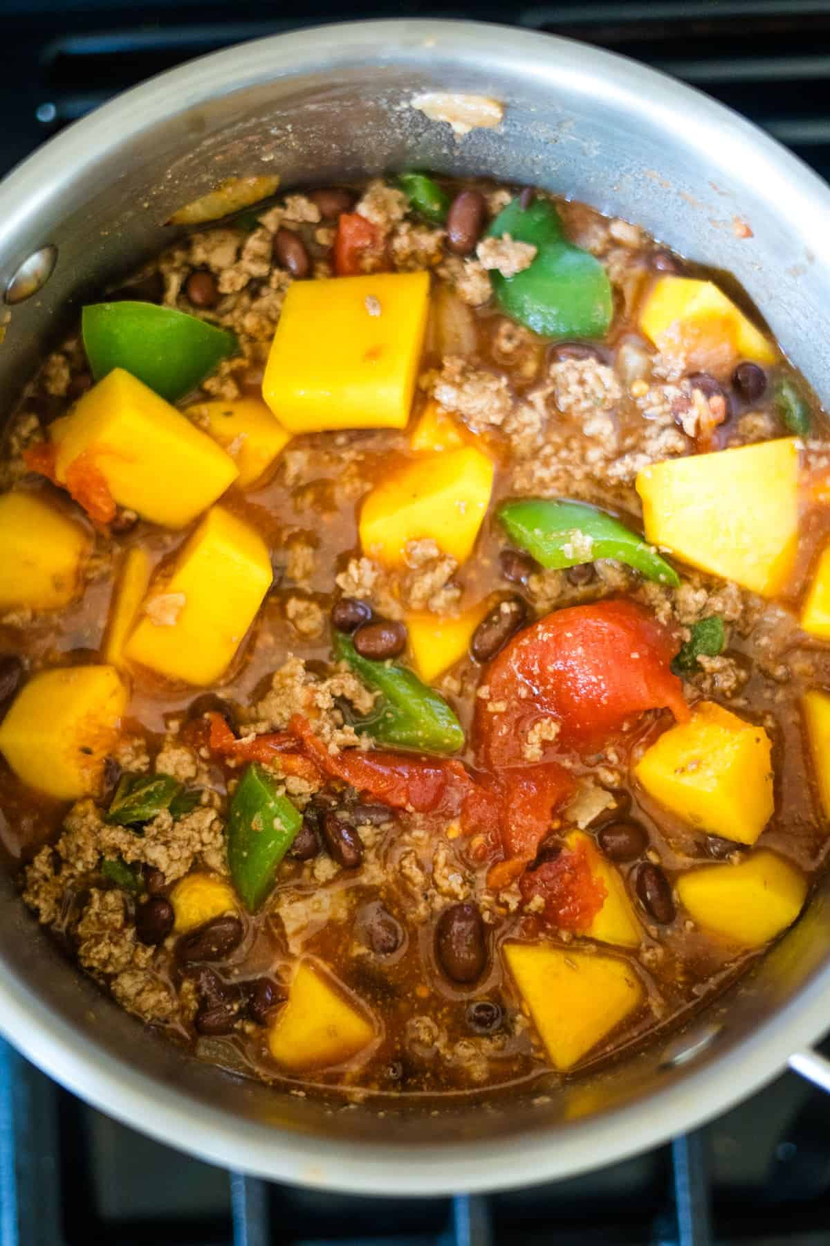 A pot filled with a hearty keto pumpkin chili containing ground meat, black beans, diced tomatoes, bell peppers, and chunks of yellow squash is bubbling on the stove.