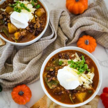 Two bowls of chili topped with sour cream, grated cheese, and chopped green onions, placed on a light-colored surface with mini pumpkins and a textured cloth.