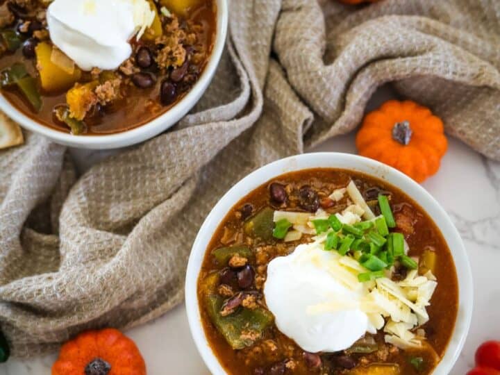 Two bowls of chili topped with sour cream, grated cheese, and chopped green onions, placed on a light-colored surface with mini pumpkins and a textured cloth.