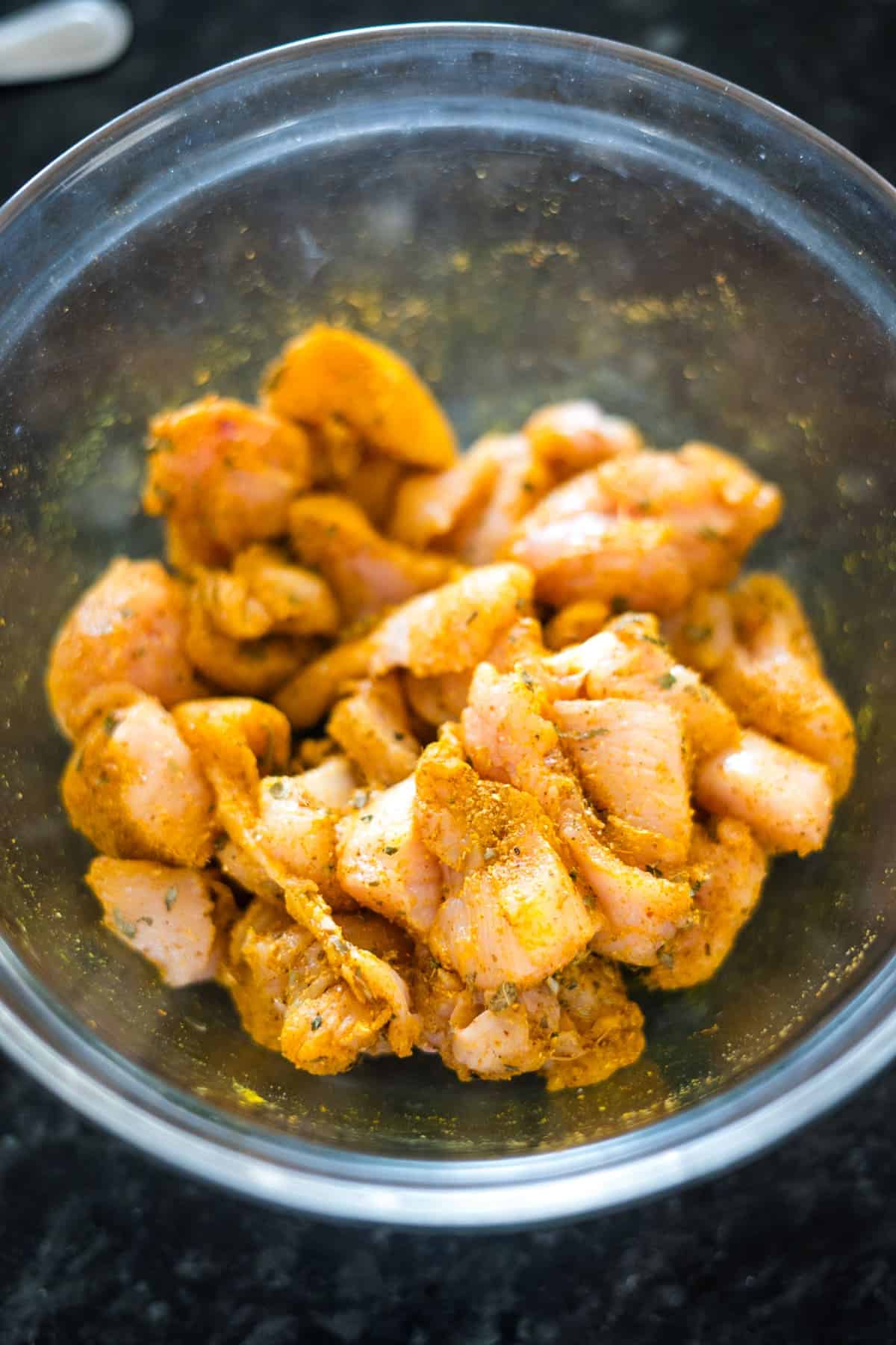 Close-up of marinated cubed chicken pieces with spices in a clear glass bowl, set against a dark background.