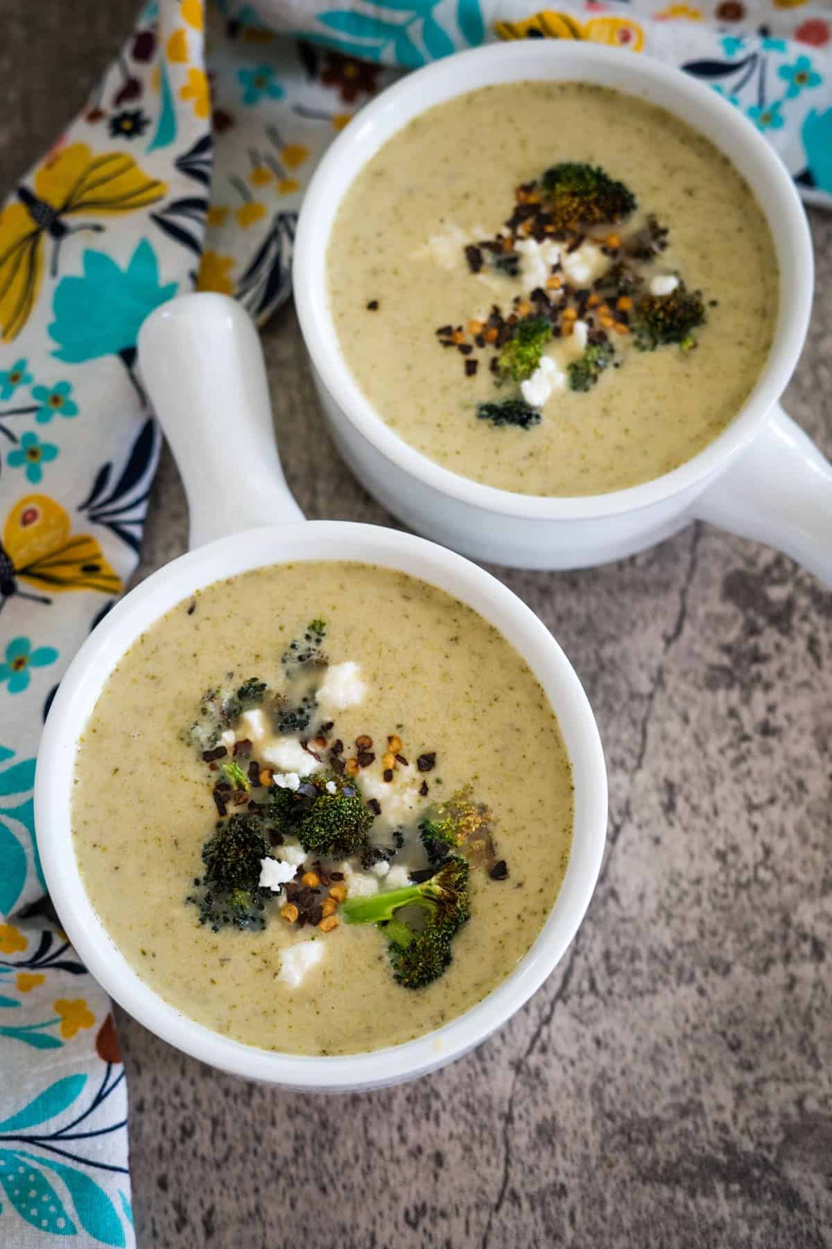 Two white bowls of broccoli soup garnished with broccoli florets and crumbled cheese, placed on a textured surface next to a colorful, floral-patterned cloth.