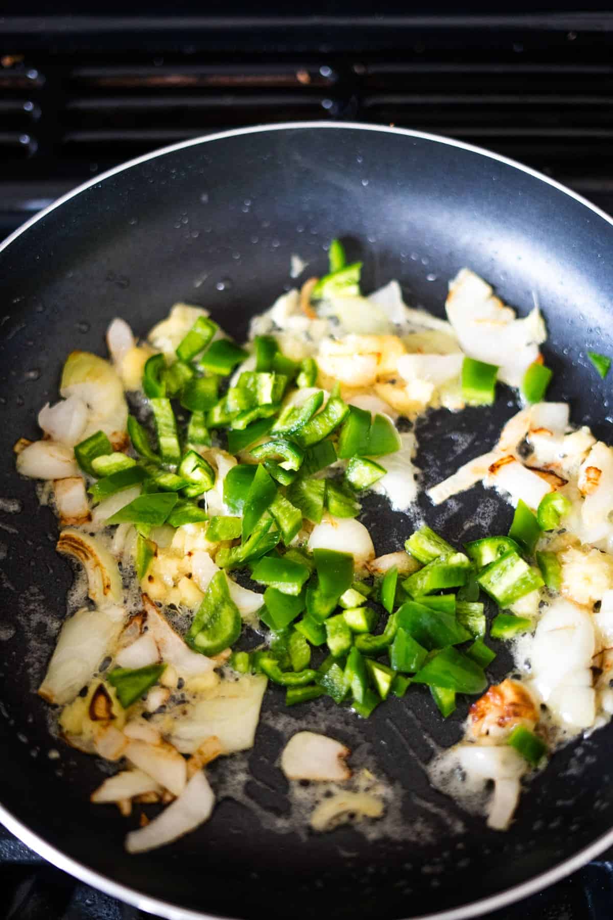 A frying pan containing chopped green bell peppers, onions, and minced garlic cooking on a stovetop.