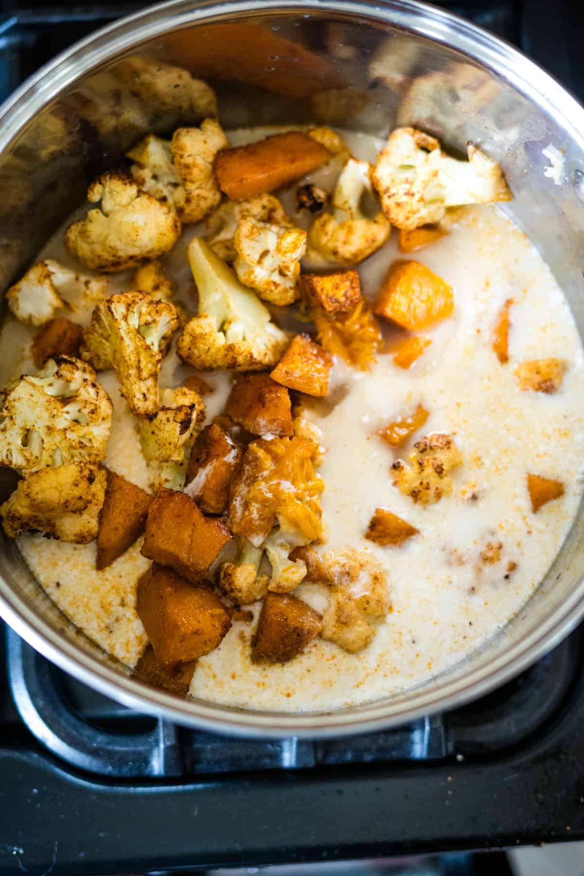 A pot on a stove containing roasted cauliflower and sweet potatoes simmering in a creamy pumpkin soup base.