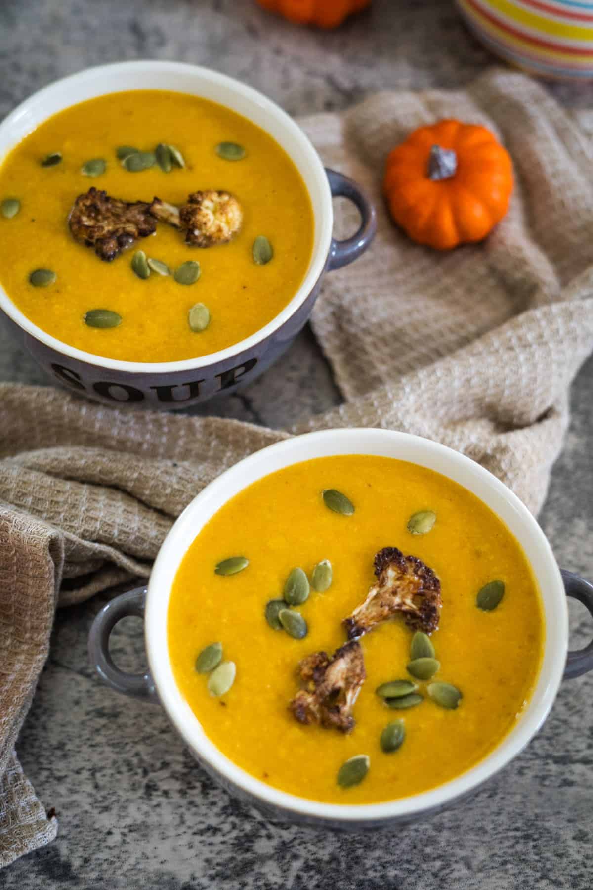 Two bowls of pumpkin cauliflower soup topped with roasted cauliflower and pumpkin seeds on a stone surface with a patterned cloth and small pumpkins in the background.