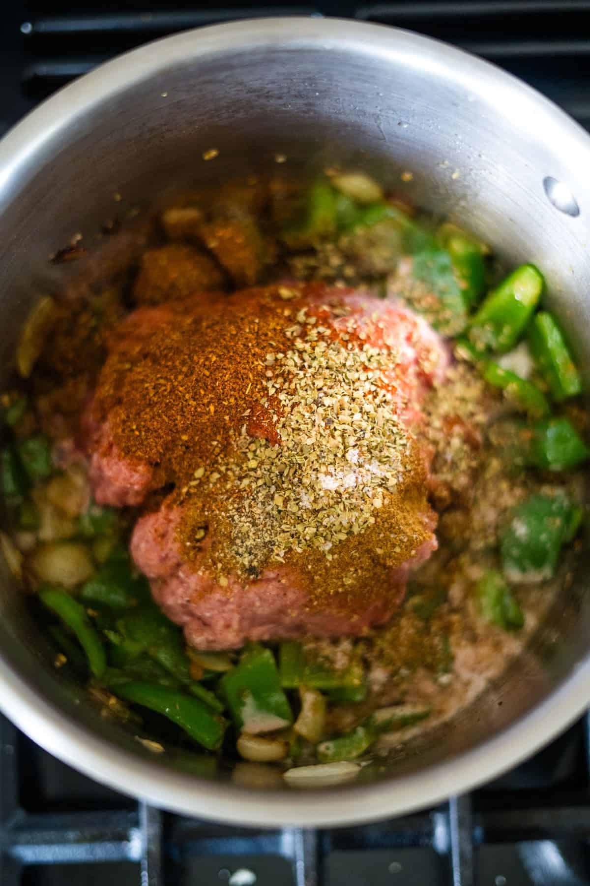 A stainless steel pot containing raw minced meat, chopped green bell peppers, onions, and various spices is ready to transform into a delicious keto pumpkin chili.