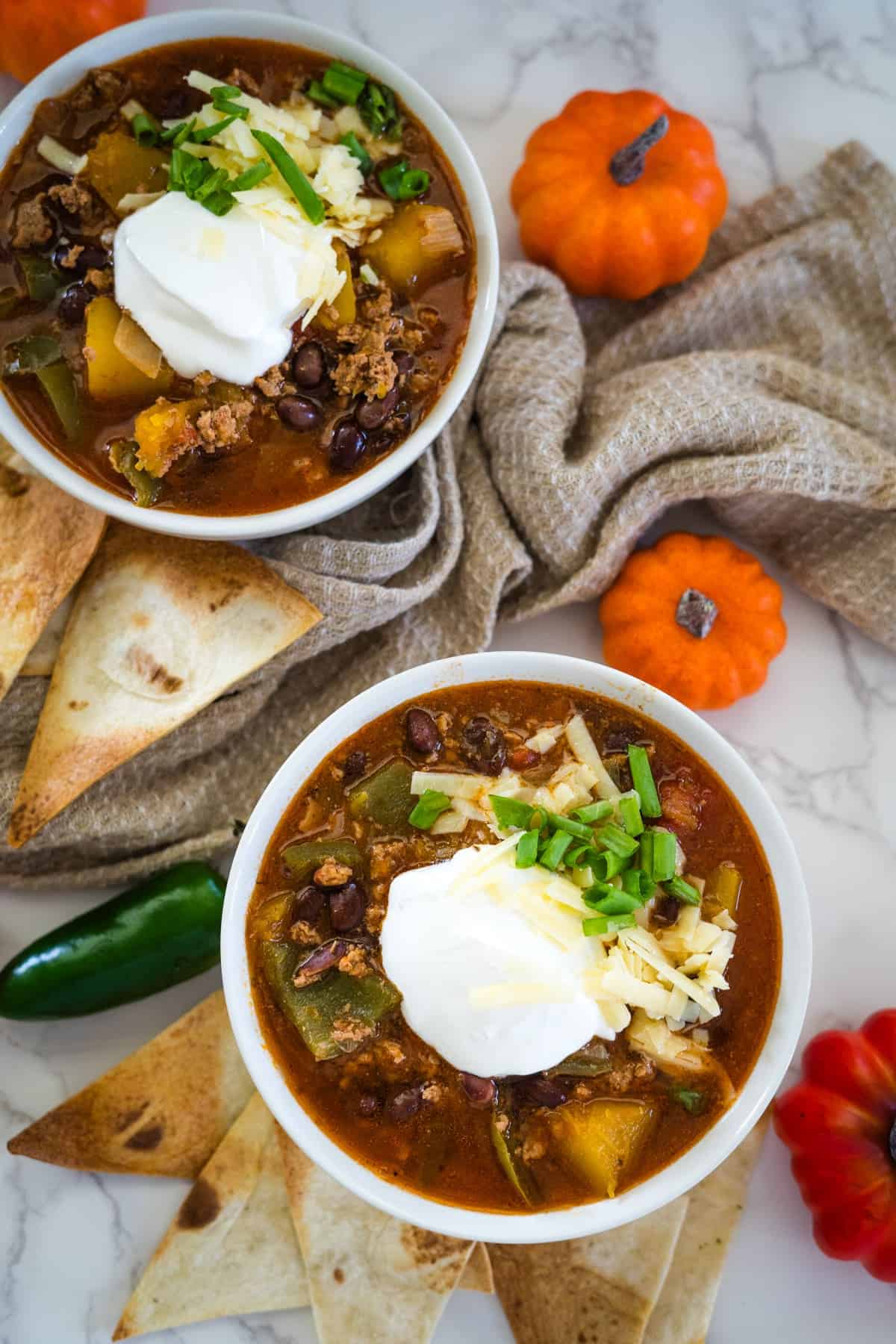 Two bowls of keto pumpkin chili topped with sour cream and shredded cheese, surrounded by tortilla chips, a jalapeño, mini pumpkins, and a beige cloth on a marble surface.