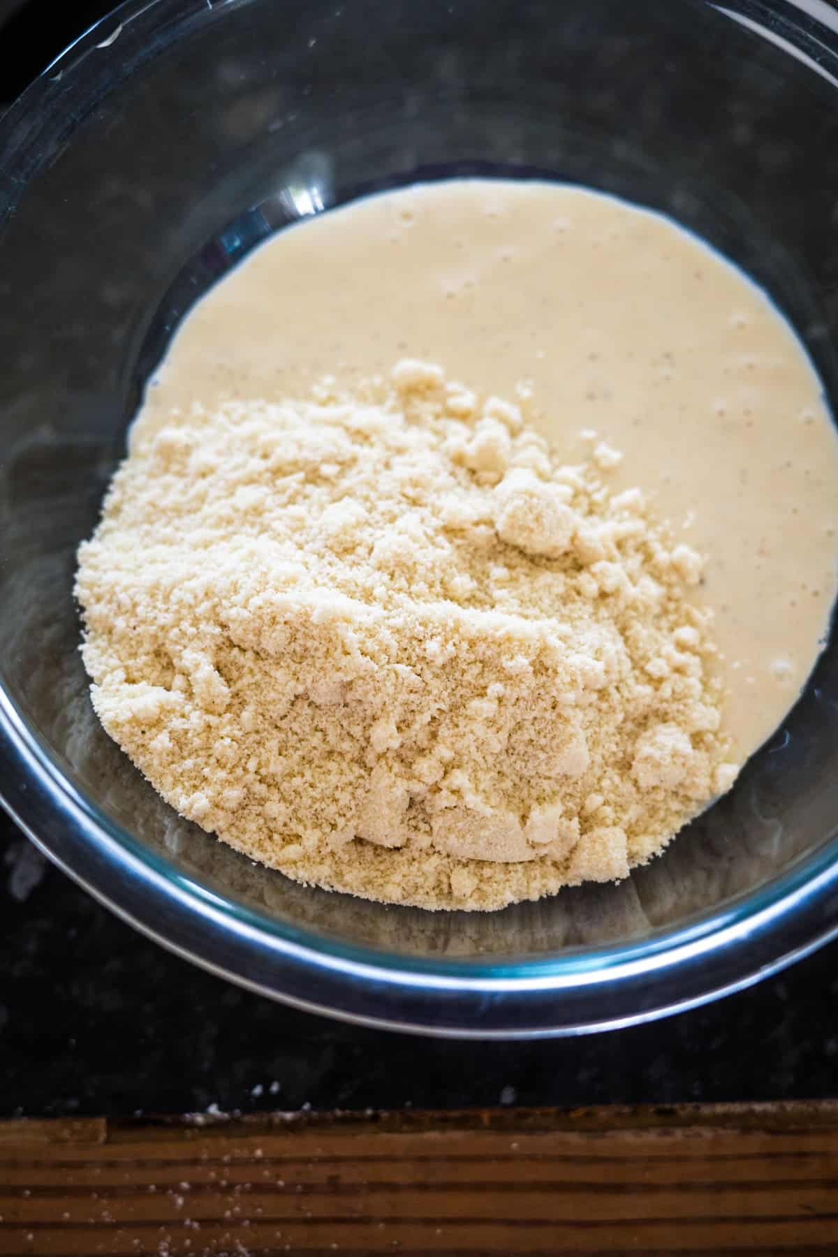 A glass bowl containing a mixture of almond flour and a liquid batter on a dark countertop.