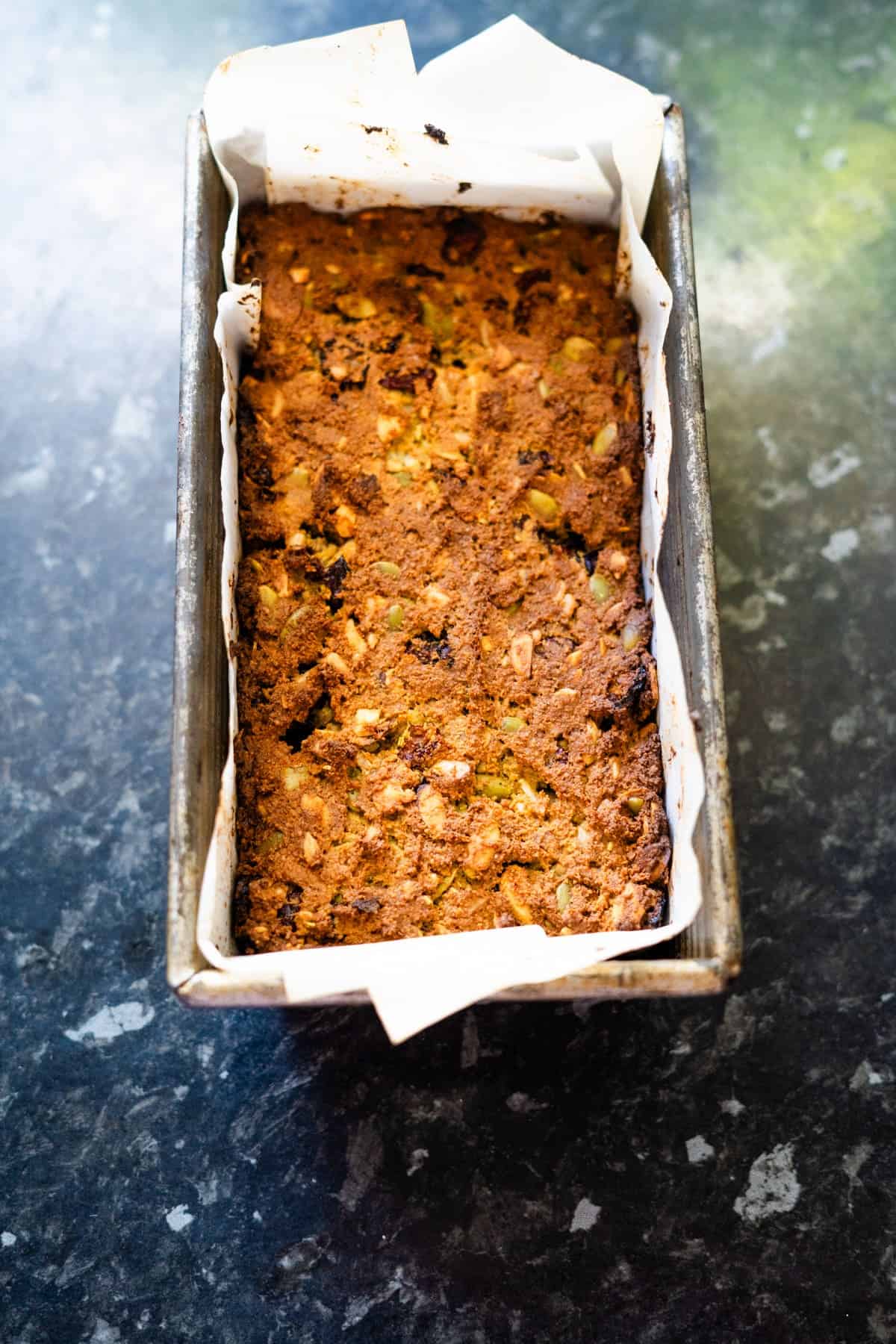 A rectangular loaf of bread with a golden crust in a parchment-lined baking pan, placed on a dark countertop.