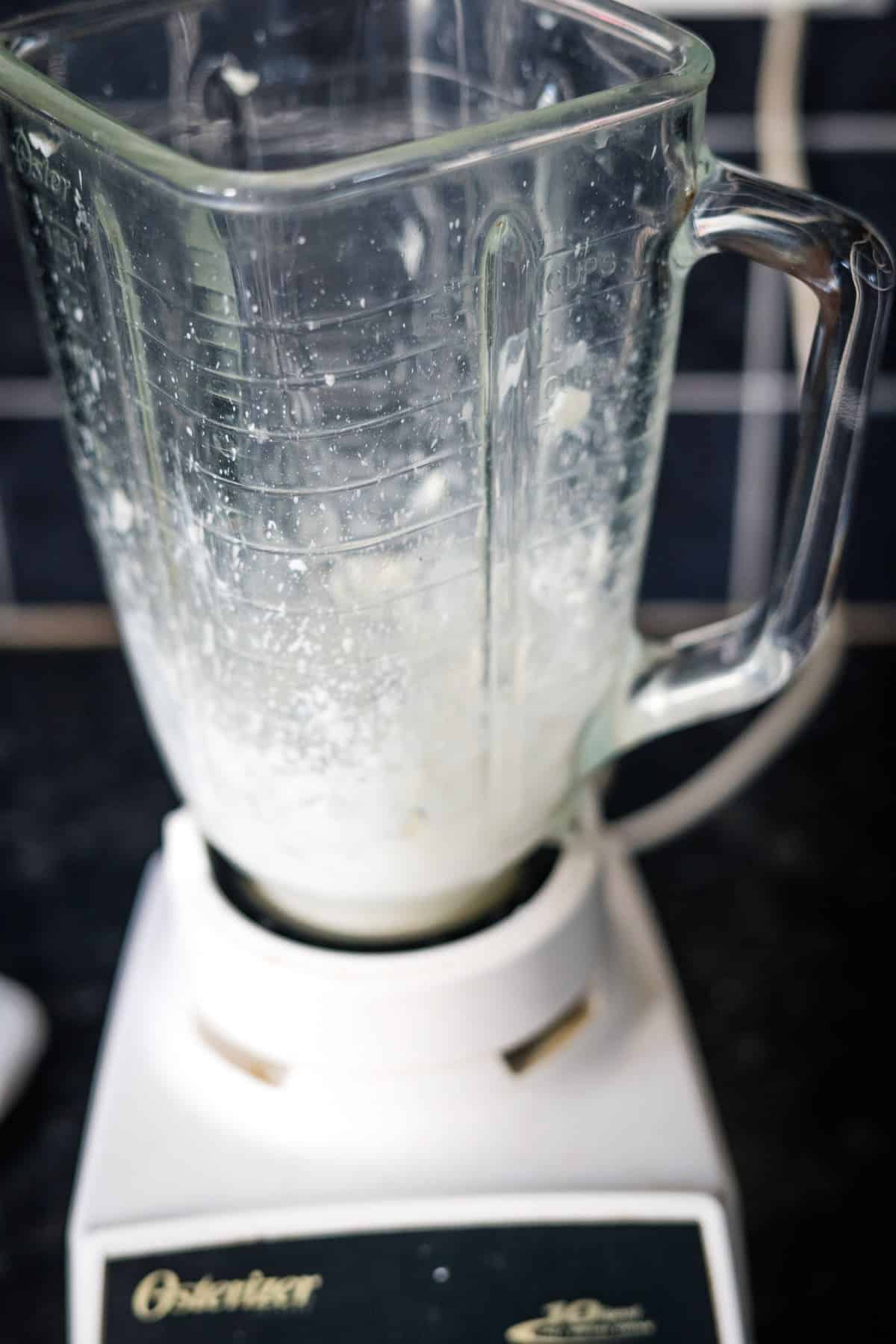 A glass blender pitcher partially filled with a white liquid, placed on a countertop.