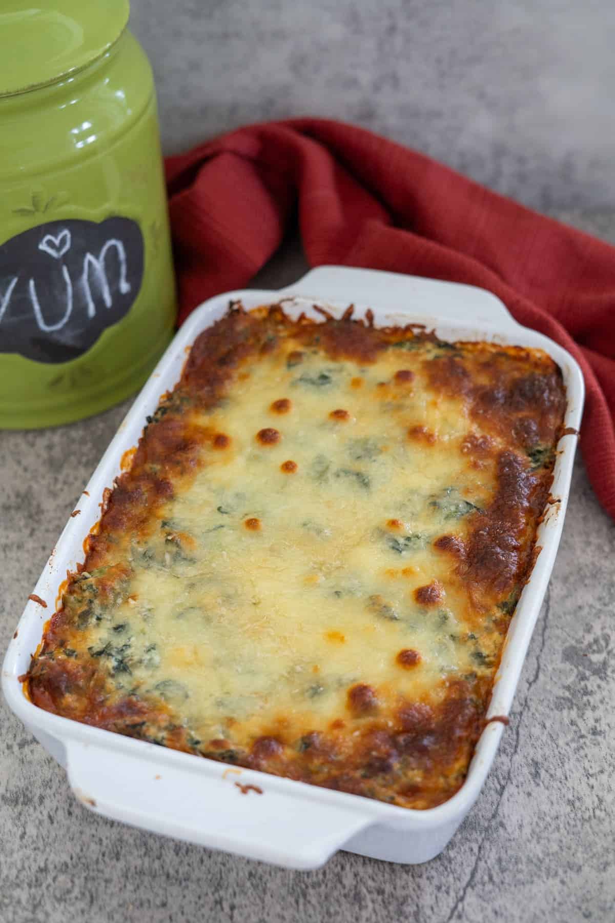 A keto spinach lasagna bakes to perfection in a rectangular white dish on a gray countertop, with a red cloth and a green container labeled "YUM" adding charm to the scene.