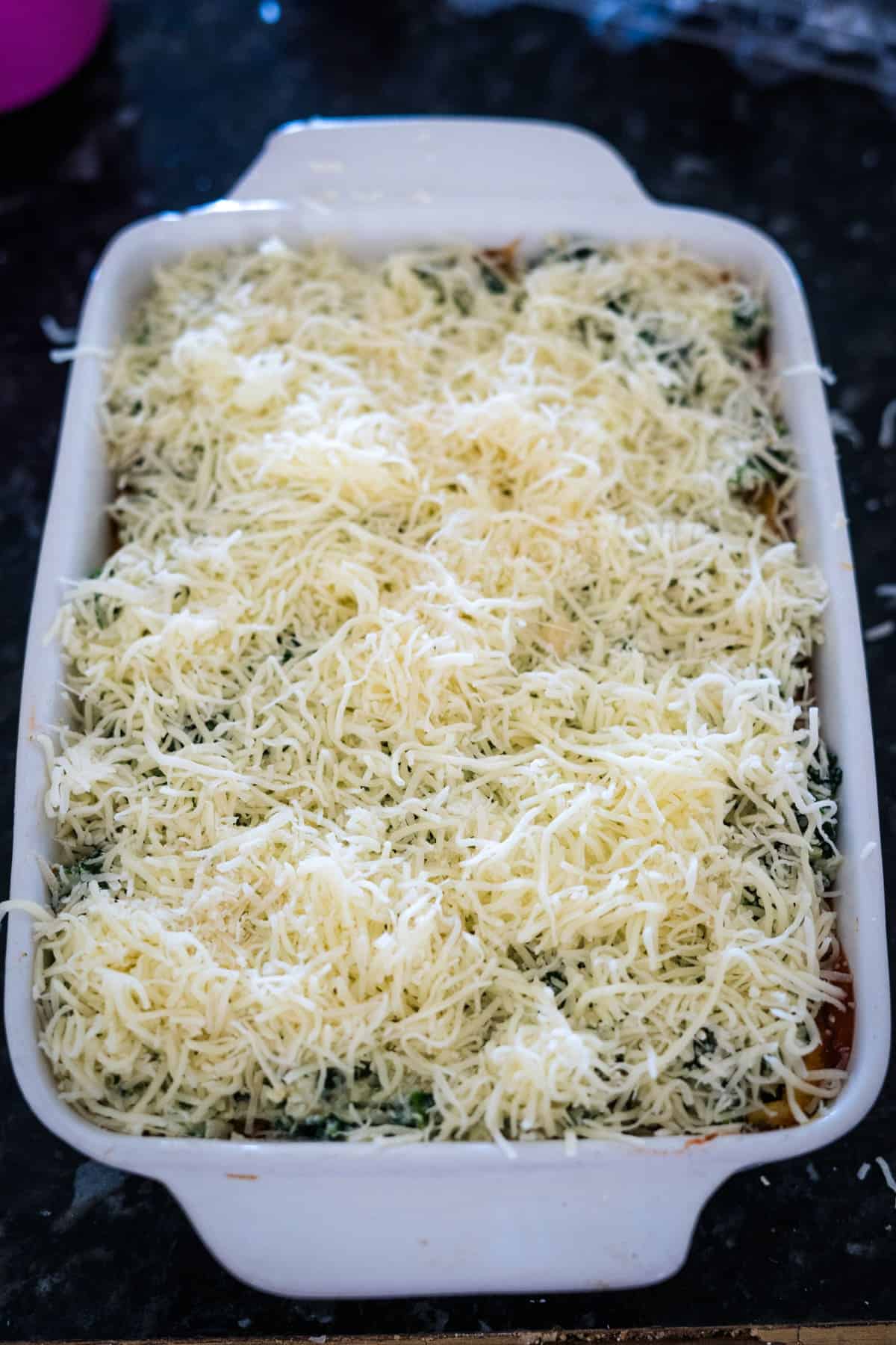 A white baking dish filled with shredded cheese on top of layered ingredients, likely prepared for baking a delicious keto spinach lasagna.