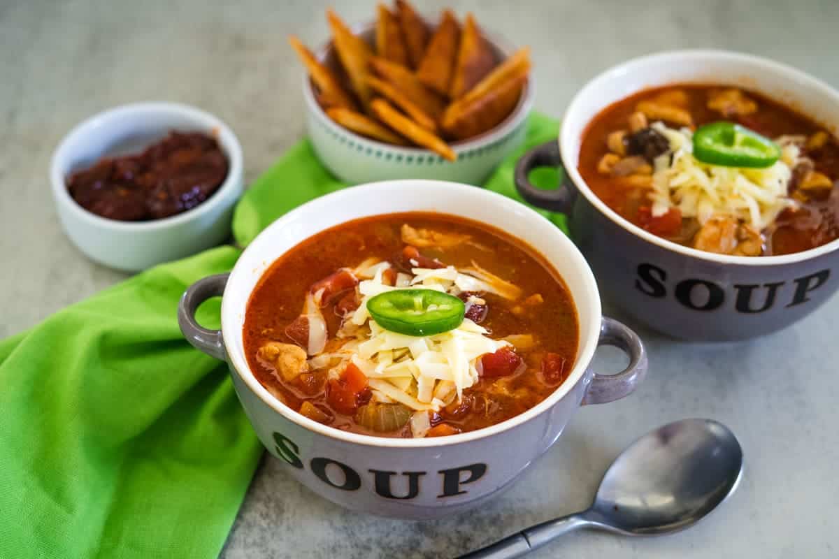 Two bowls of soup, labeled "Soup," brimming with shredded cheese and jalapeño, suggest a hint of chicken chipotle flavor. Next to them, a spoon awaits, while a small bowl of sauce and a dish with potato wedges linger in the background on a green cloth.