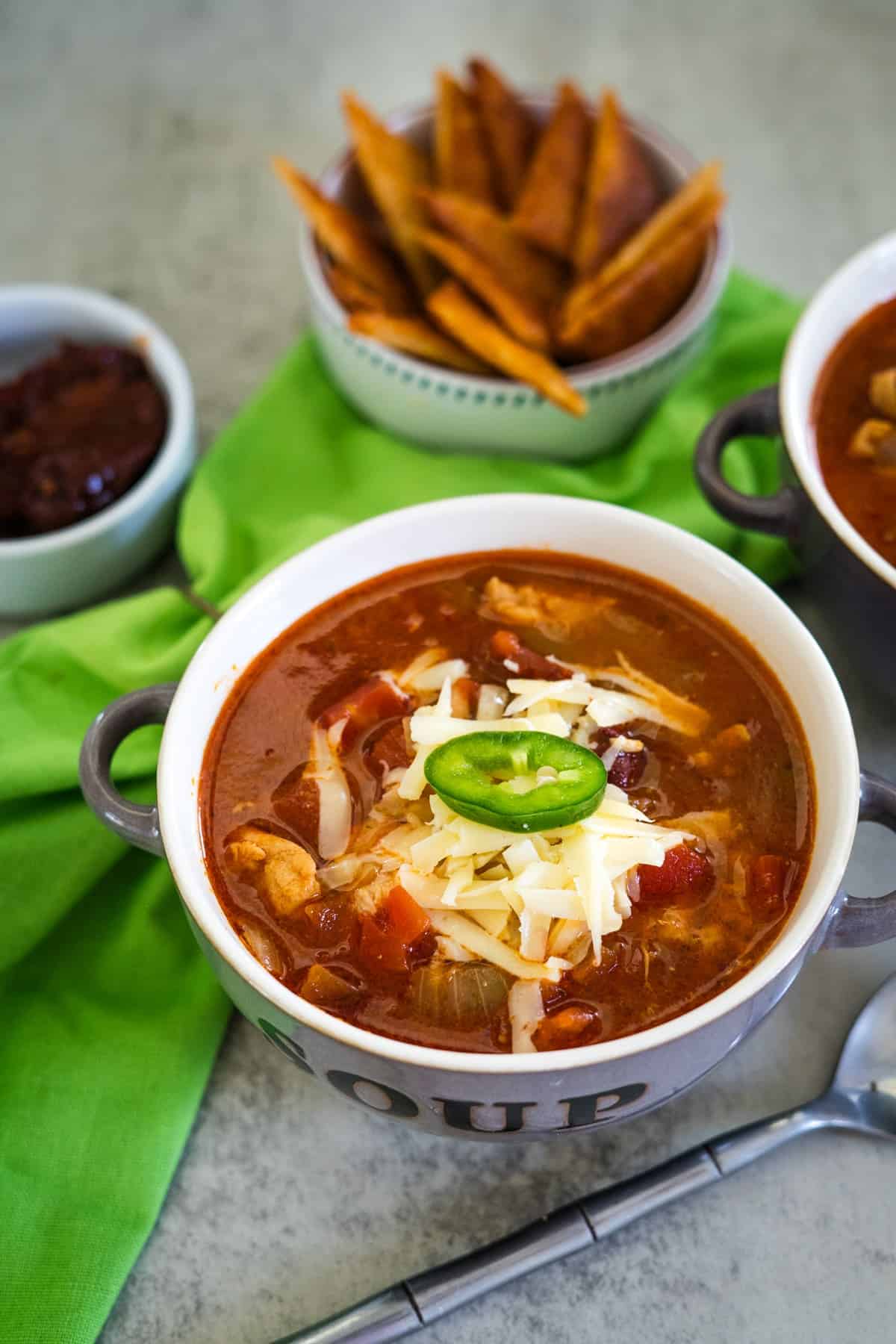 A bowl of chili topped with cheese and a jalapeño slice, accompanied by crispy wedges and a small dish of sauce, all resting invitingly on a green cloth. This savory scene hints at a hearty chicken chipotle soup waiting in the wings.