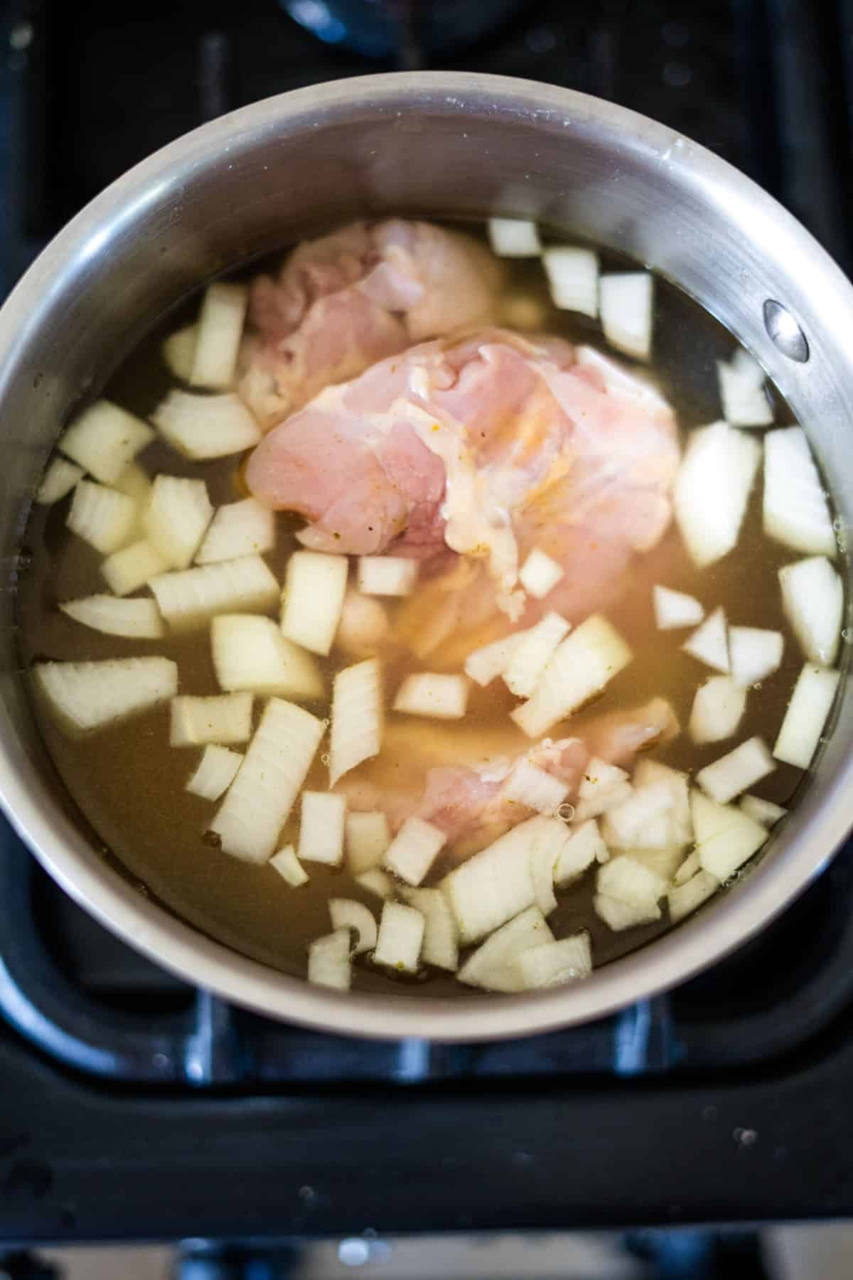 Pot with raw chicken pieces and chopped onions in water on a stove.