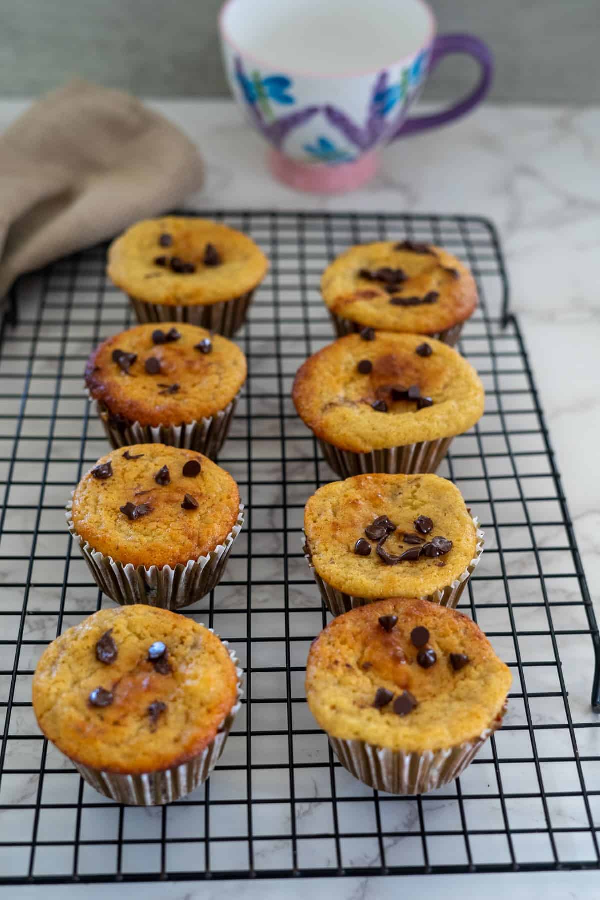 Nine muffins with chocolate chips cooling on a wire rack, accompanied by a purple-handled mug in the background.