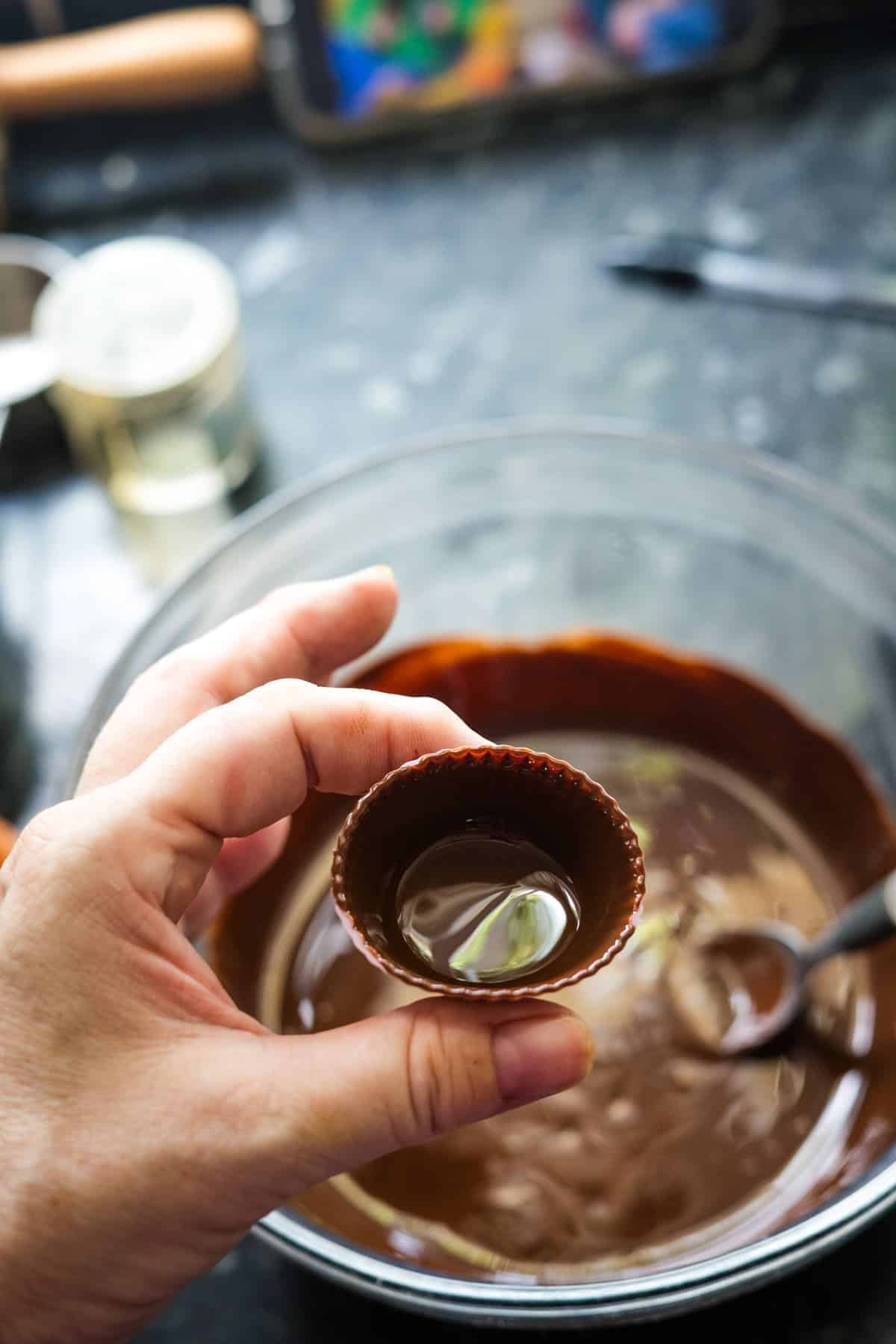 A hand holding a small, empty chocolate cup over a bowl of melted chocolate with a spoon inside, on a dark kitchen countertop, hints at crafting delectable high-protein peanut butter cups.