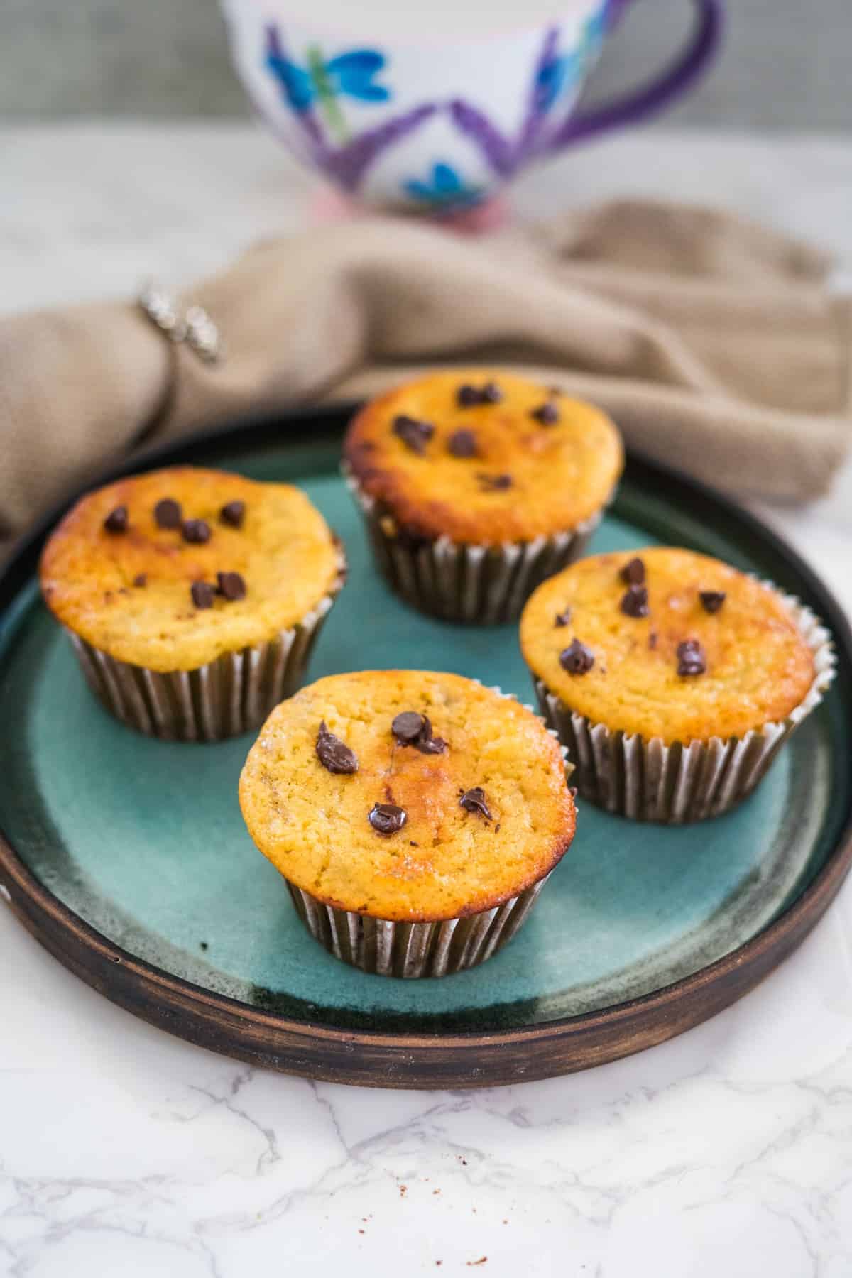 Four cottage cheese chocolate chip muffins rest on a green plate, accompanied by a cup and beige cloth in the background.