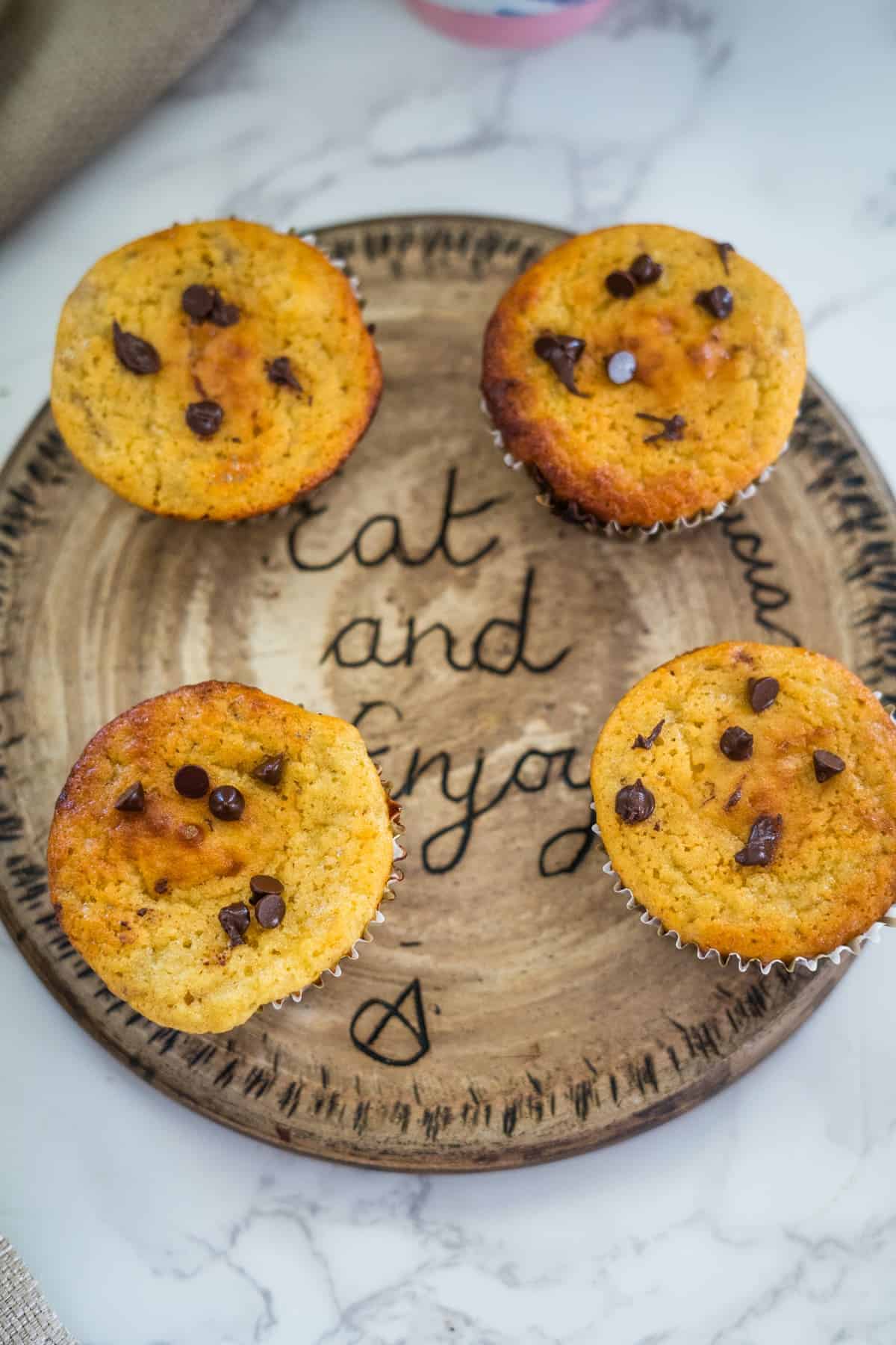 Four delightful cottage cheese chocolate chip muffins rest on a round wooden plate with "Eat and Enjoy" elegantly inscribed.