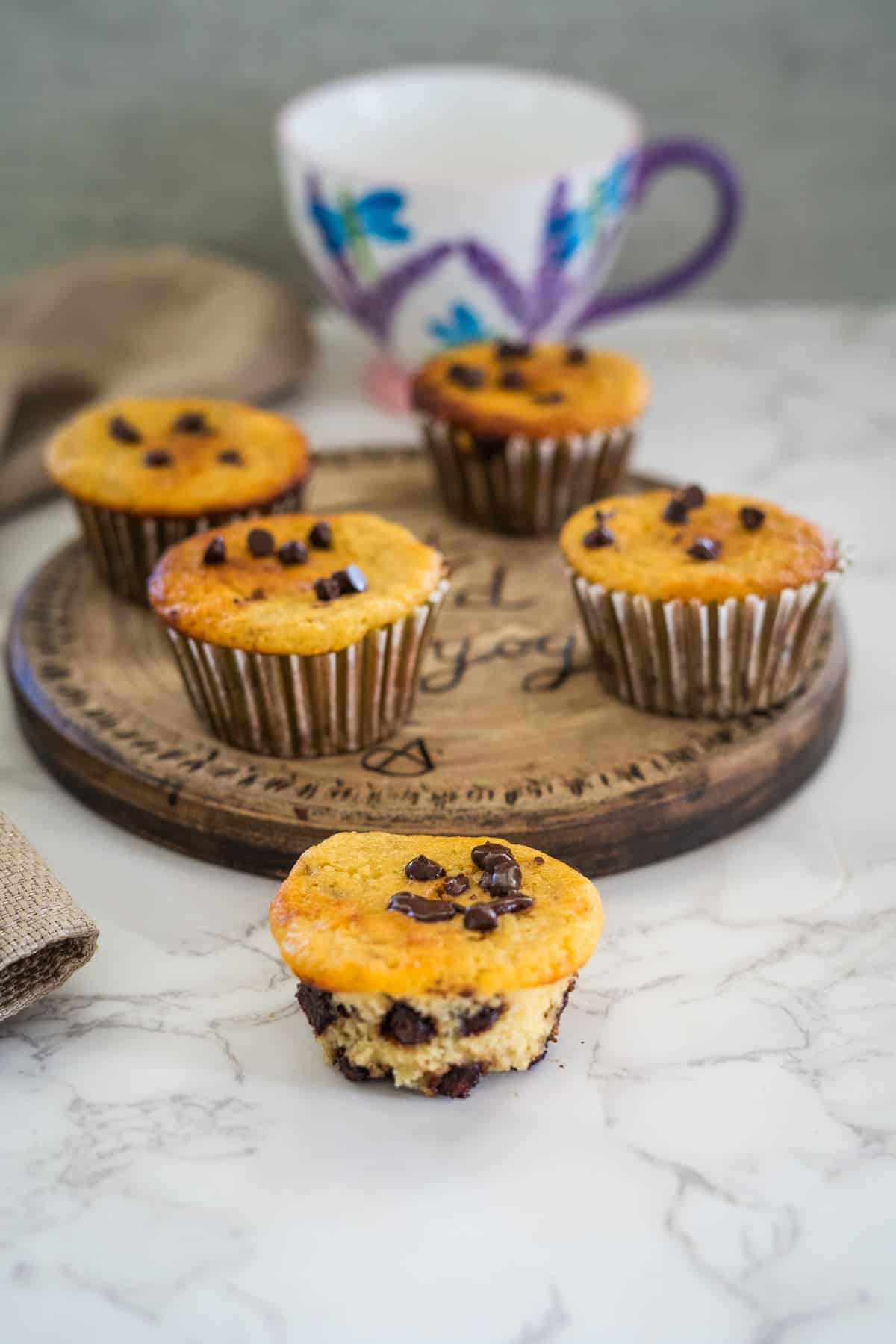 Six chocolate chip muffins on a wooden platter with one muffin in the foreground. A decorative mug is in the background.