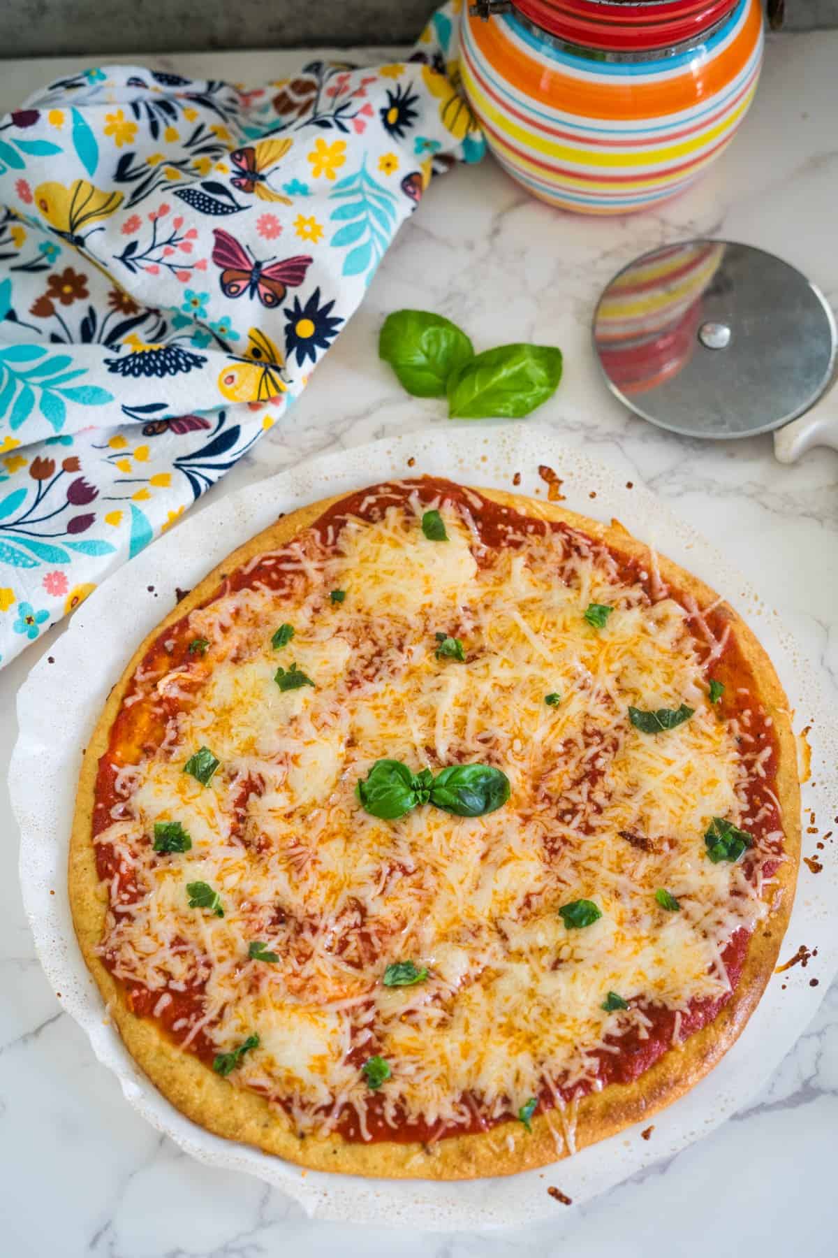 A freshly baked pizza topped with melted cheese and herbs on a marble countertop, accompanied by a pizza cutter and a colorful fabric.