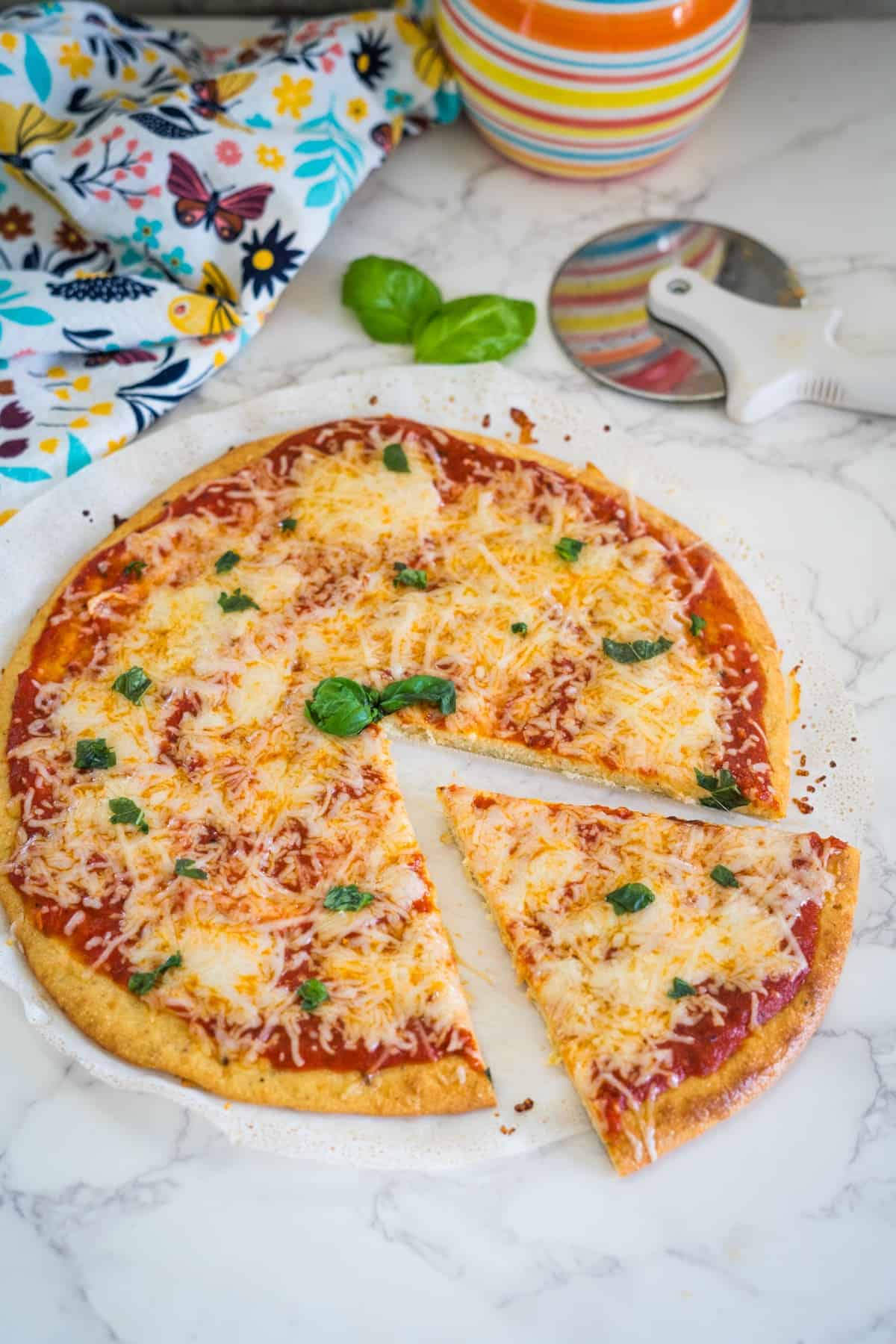 A cheese pizza on a marble countertop with one slice cut out. A pizza cutter, colorful mug, and floral cloth are nearby. Basil leaves garnish the pizza.