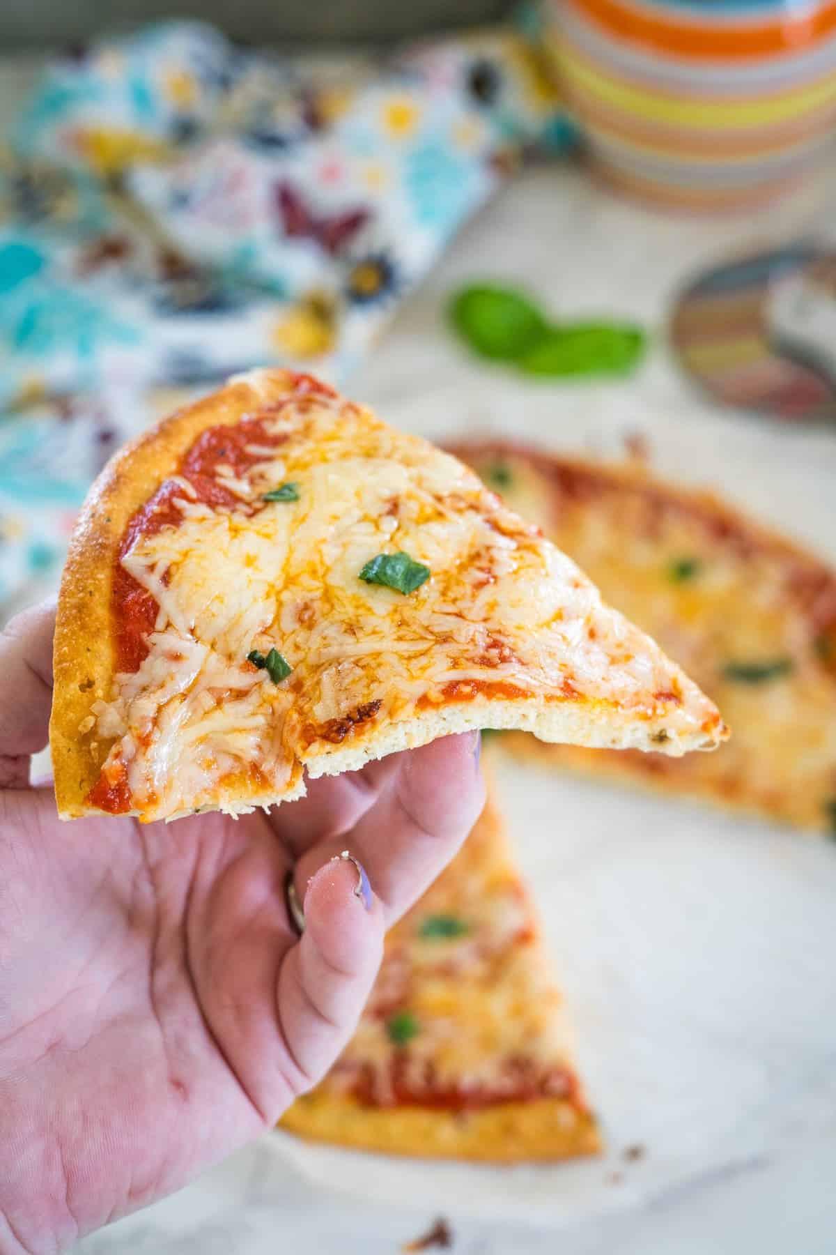 A hand holding a slice of cheese pizza with basil, with the rest of the pizza in the background on a white surface.