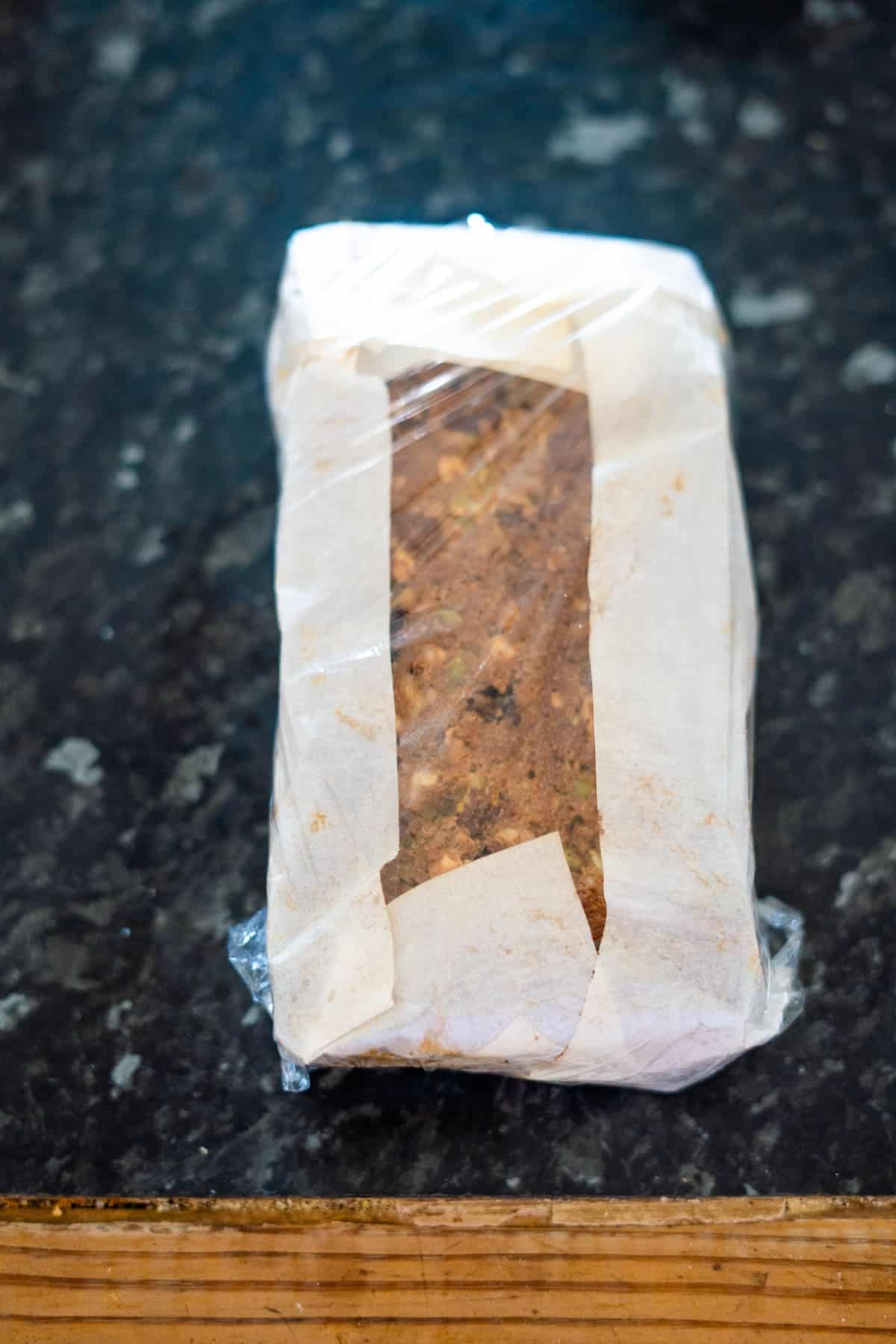 A loaf of bread wrapped in clear plastic and parchment paper on a dark marble countertop.