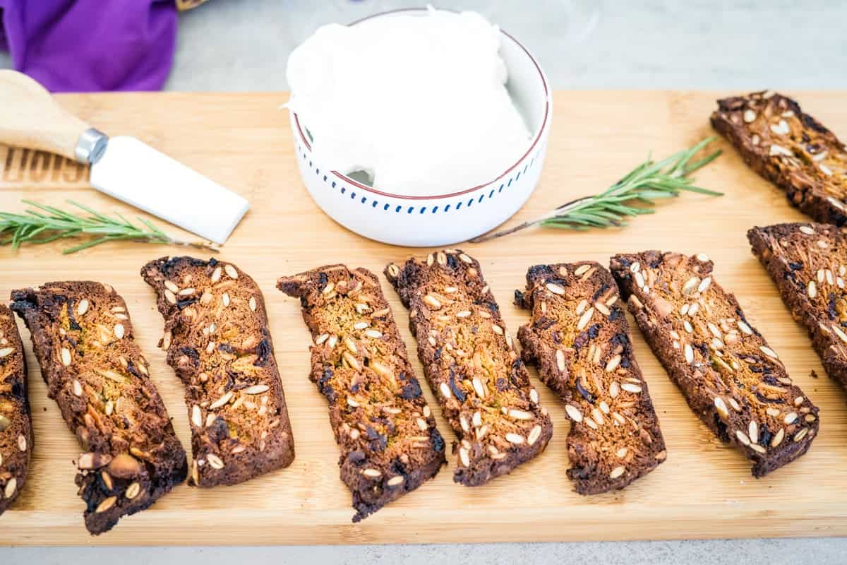 Slices of seeded bread are arranged on a wooden board next to a bowl of white spread and a butter knife. Rosemary sprigs are placed nearby.