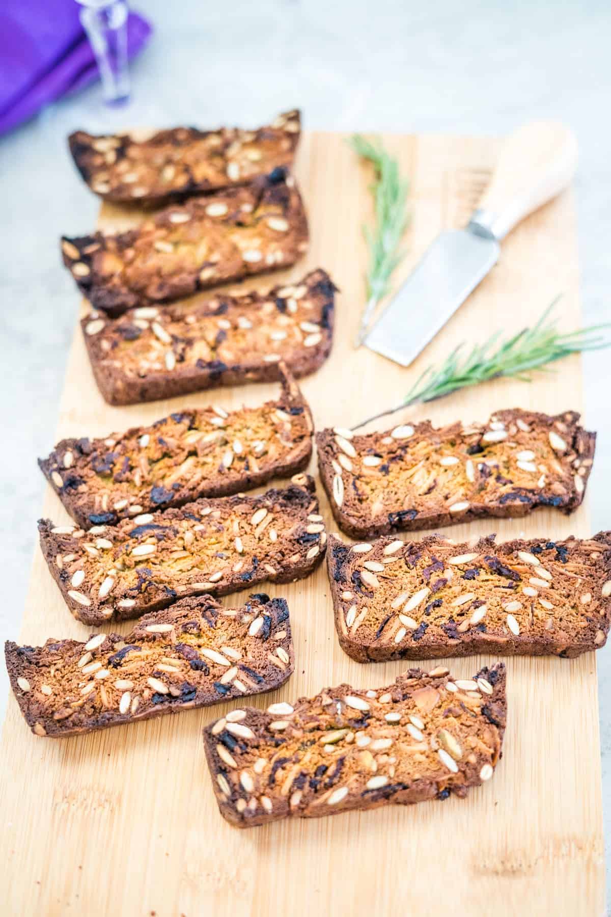 Sliced crispy bread with seeds and herbs on a wooden board next to a knife and sprigs of rosemary.