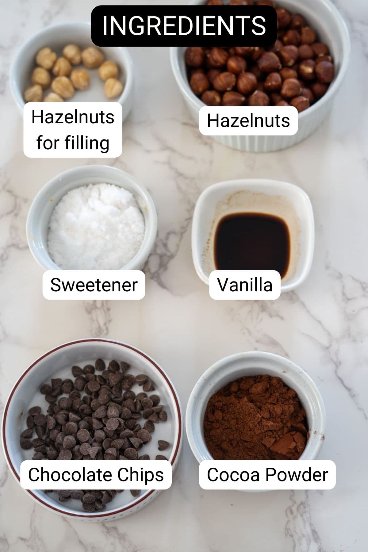 A top-down view of various labeled ingredients in bowls on a marble surface: hazelnuts for filling, hazelnuts, sweetener, vanilla, chocolate chips, and cocoa powder for a sugar-free Ferrero Rocher.