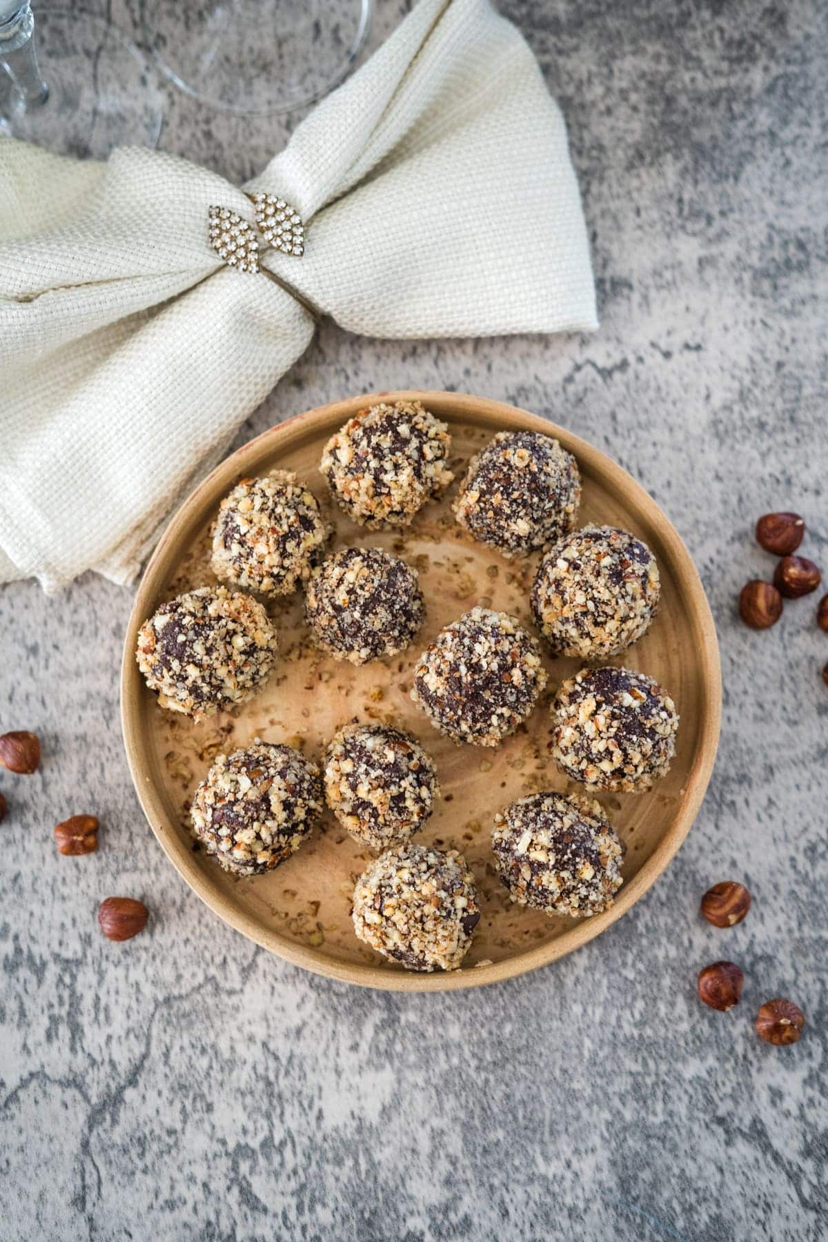 A wooden plate with round chocolate truffles coated in chopped nuts, placed on a gray textured surface. Scattered nuts surround the plate, and a white fabric with a jeweled brooch is in the background.