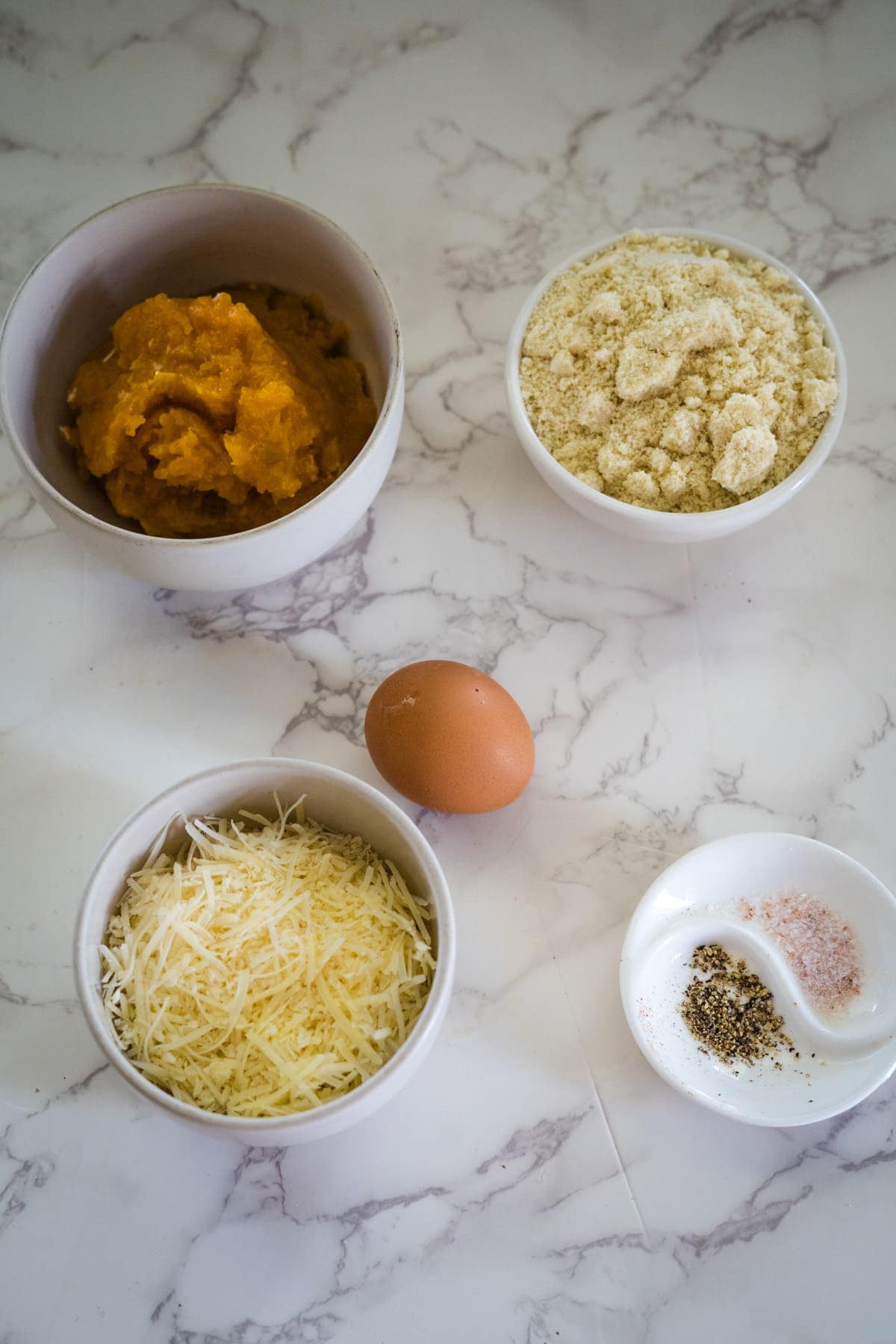 Ingredients for gluten-free pumpkin gnocchi on a marble surface: bowl of pumpkin puree, bowl of almond flour, grated cheese, one brown egg, and a small dish with pink salt and black pepper.