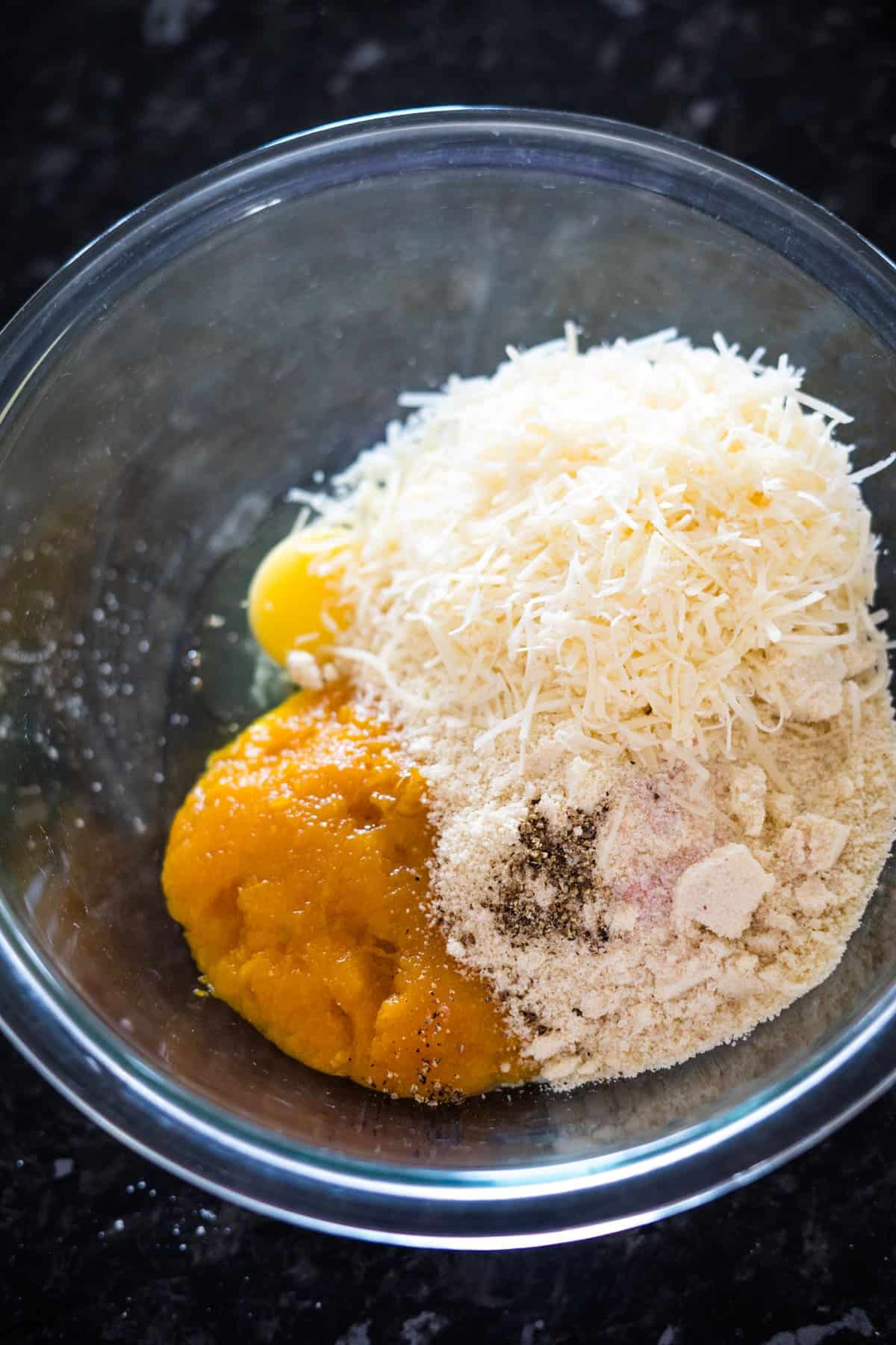 A glass bowl contains pumpkin puree, grated cheese, flour, an egg, and seasoning, ready to be mixed.