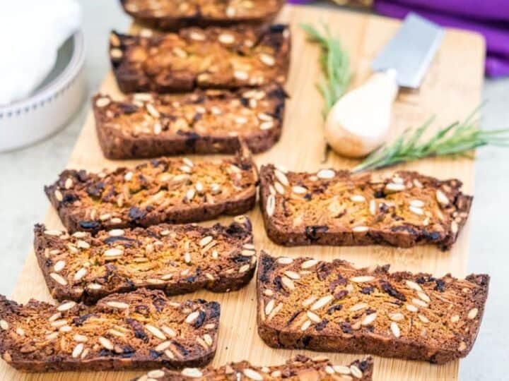 Slices of nut-and-seed bread arranged on a wooden board with a sprig of rosemary and a small cheese knife nearby.