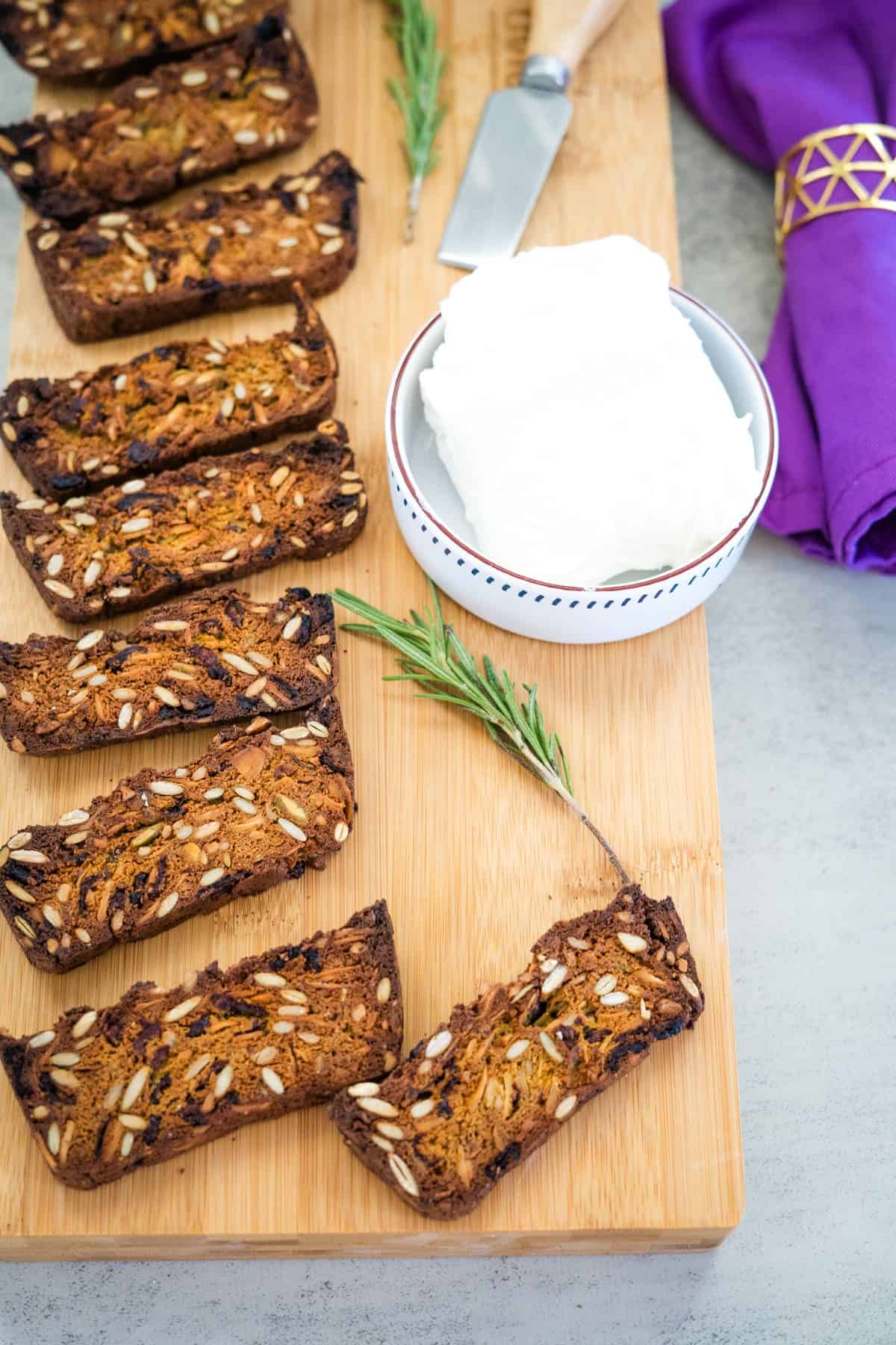 Several slices of multigrain bread with seeds on a wooden board, next to a bowl of cream cheese, a sprig of rosemary, a butter knife, and a purple napkin.