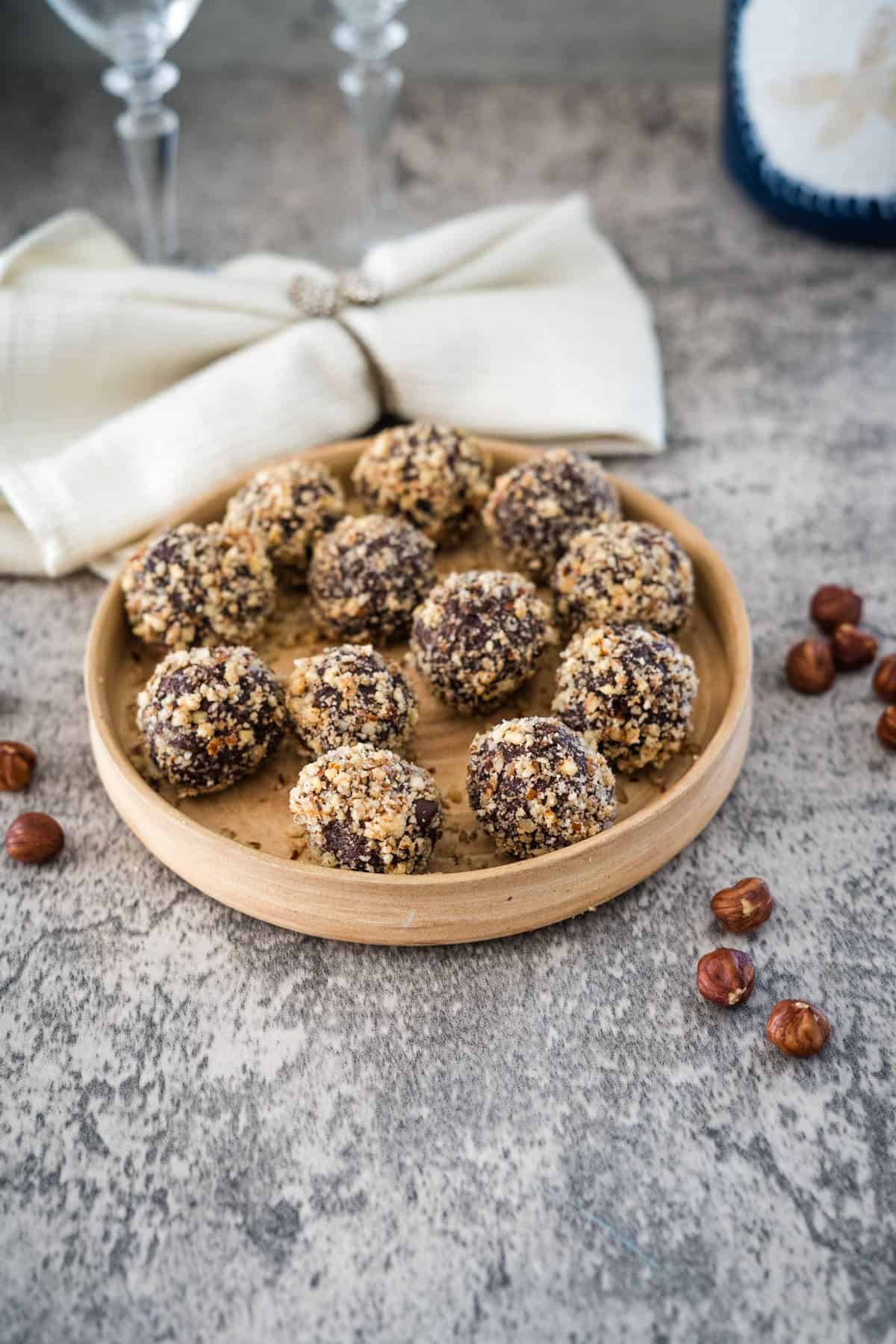 A wooden plate holds sugar-free Ferrero Rocher truffles covered in chopped nuts. Scattered hazelnuts surround the plate on a textured grey surface with a white cloth and two wine glasses in the background.