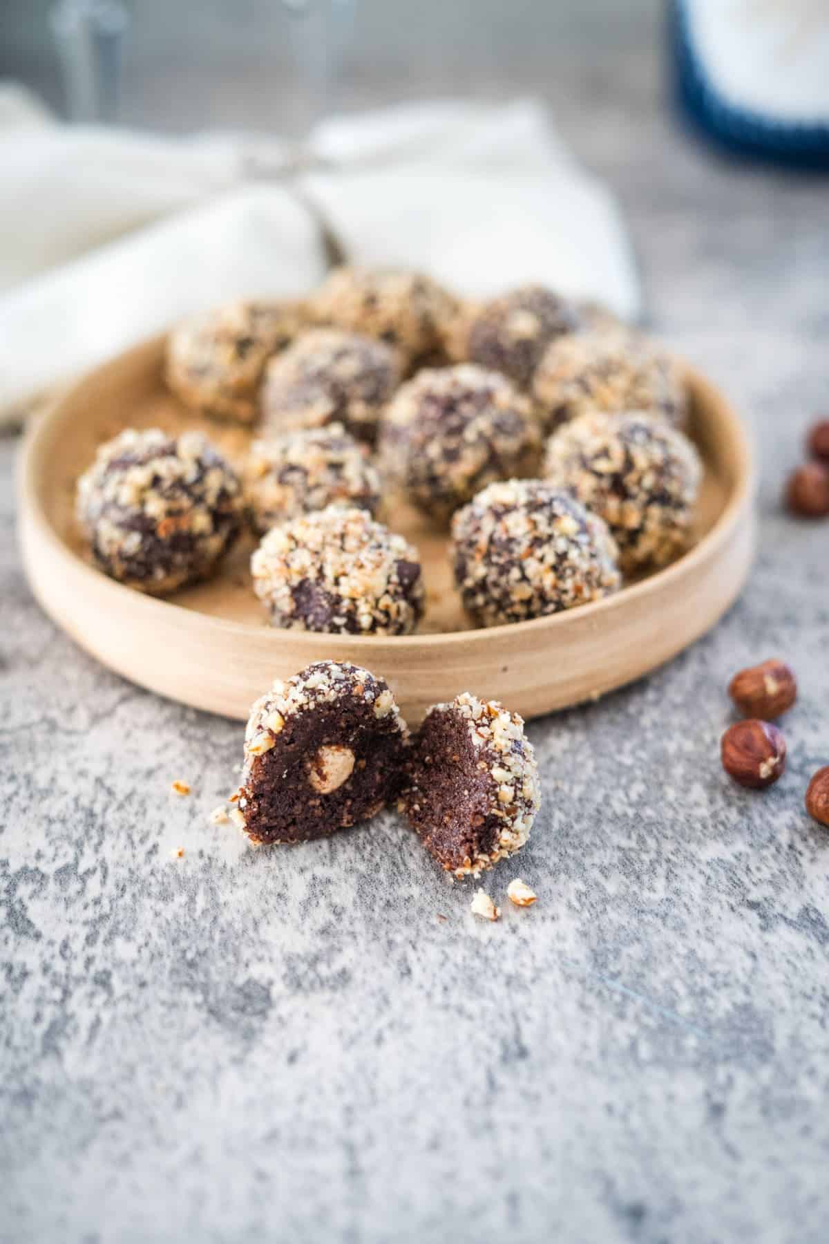 A plate of sugar-free chocolate truffles coated in crushed nuts sits on a gray surface, with one truffle cut open to reveal a whole hazelnut inside. Resembling sugar-free Ferrero Rocher, several whole hazelnuts are scattered nearby.