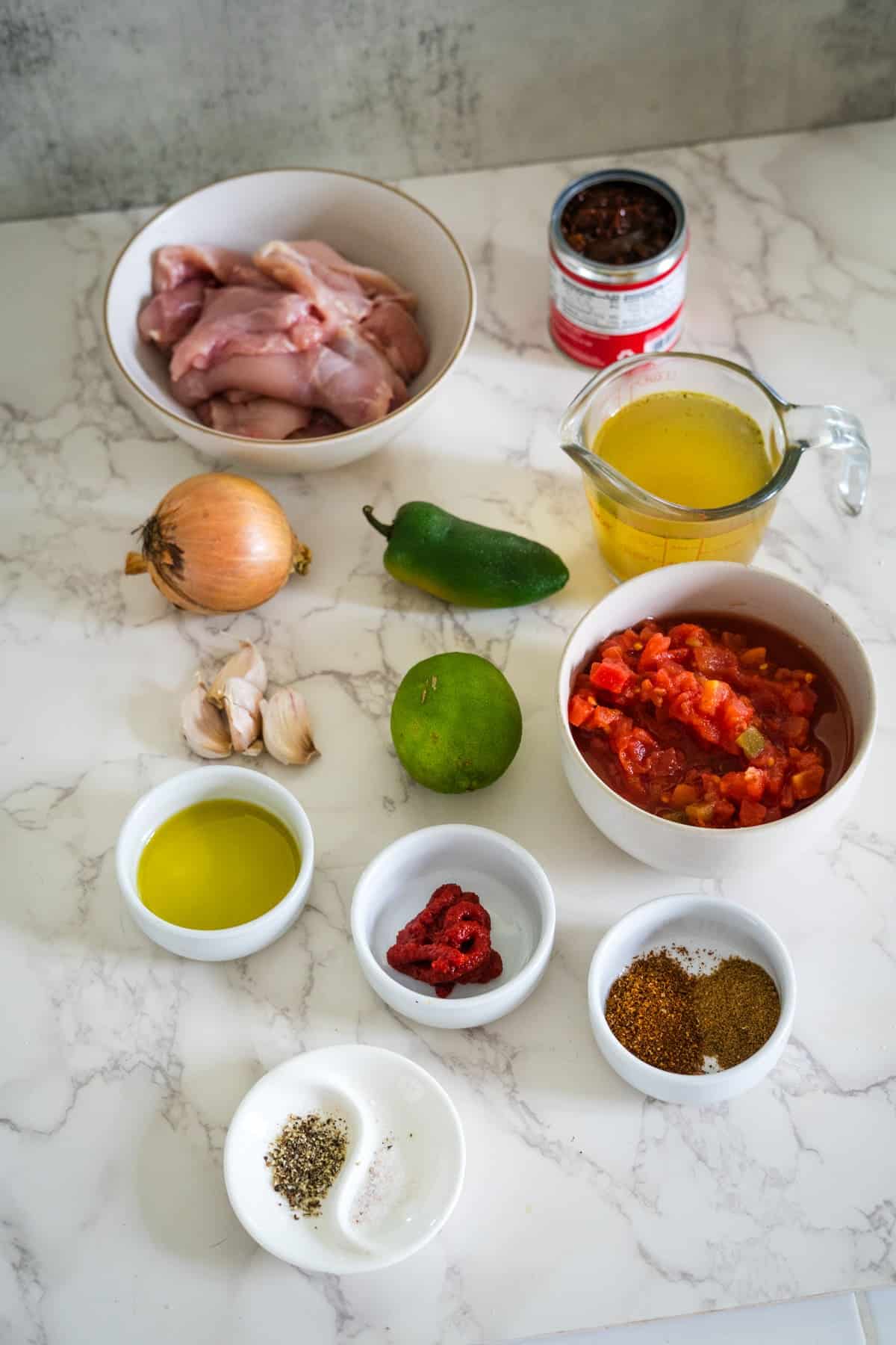 Ingredients for a dish displayed on a countertop: chicken, canned tomatoes, broth, onion, lime, jalapeño, garlic, olive oil, spices, and tomato paste.