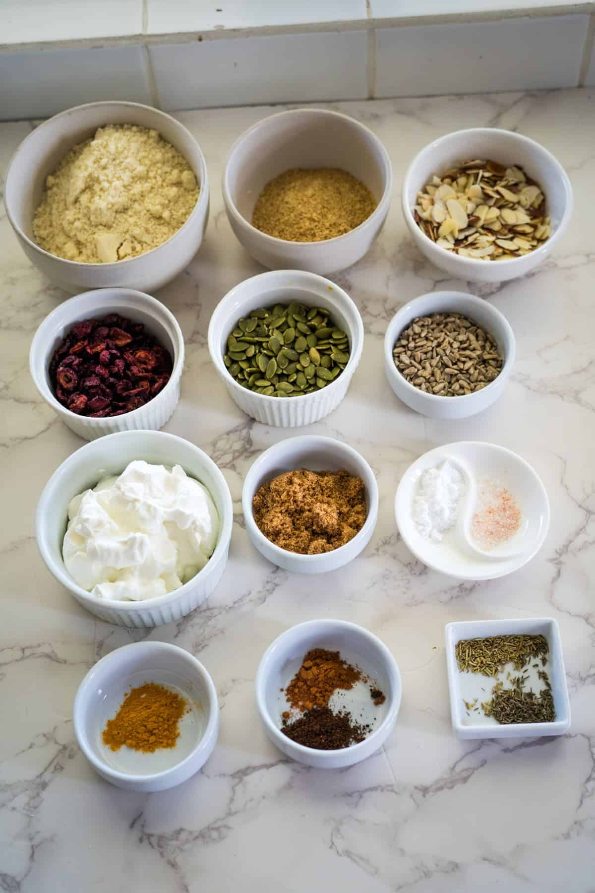 Various bowls on a countertop contain almond flour, flaxseed meal, sliced almonds, cranberries, pumpkin seeds, sunflower seeds, sour cream, brown sugar, spices, and seasoning.