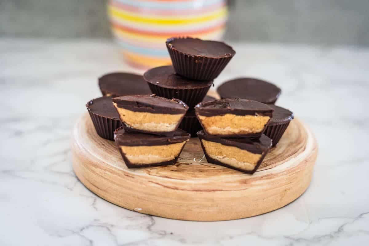 A stack of high protein chocolate peanut butter cups is displayed on a wooden board, set on a marble surface. A colorful striped container is in the blurred background.