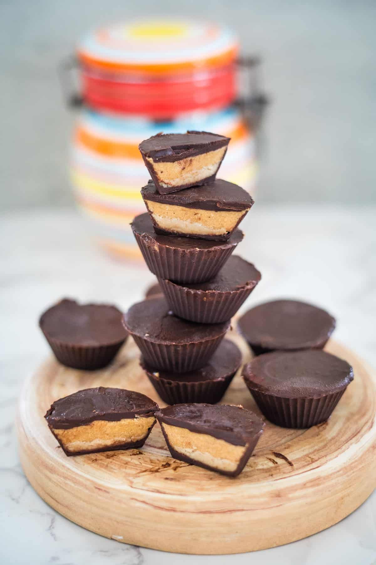 A stack of chocolate peanut butter cups on a wooden board, with a colorful jar in the background.