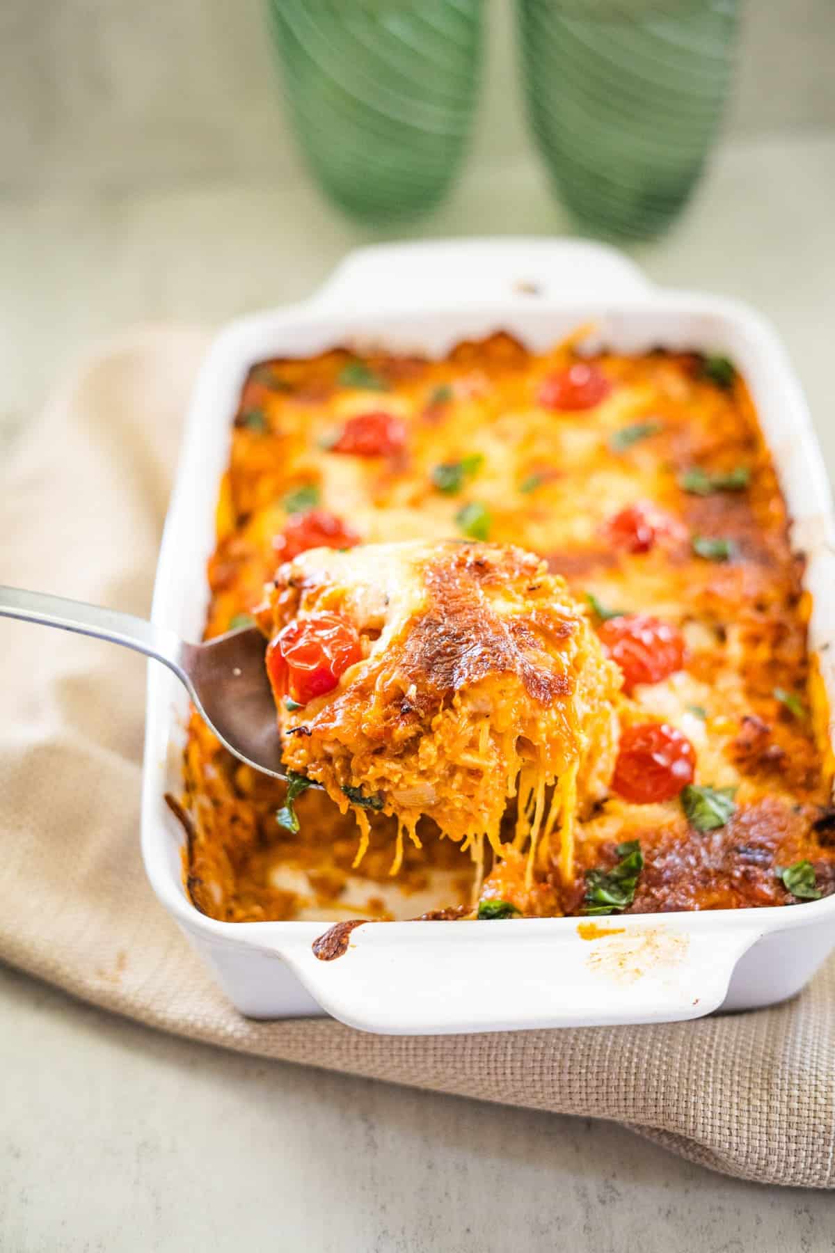 A serving of baked spaghetti squash pizza casserole with cheese and cherry tomatoes is lifted from a white baking dish.