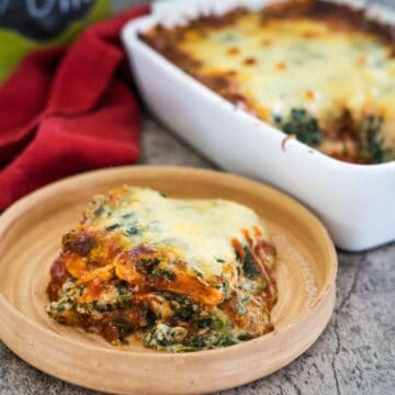 A slice of vegetable lasagna with melted cheese on a wooden plate, next to a baking dish containing the rest of the lasagna.