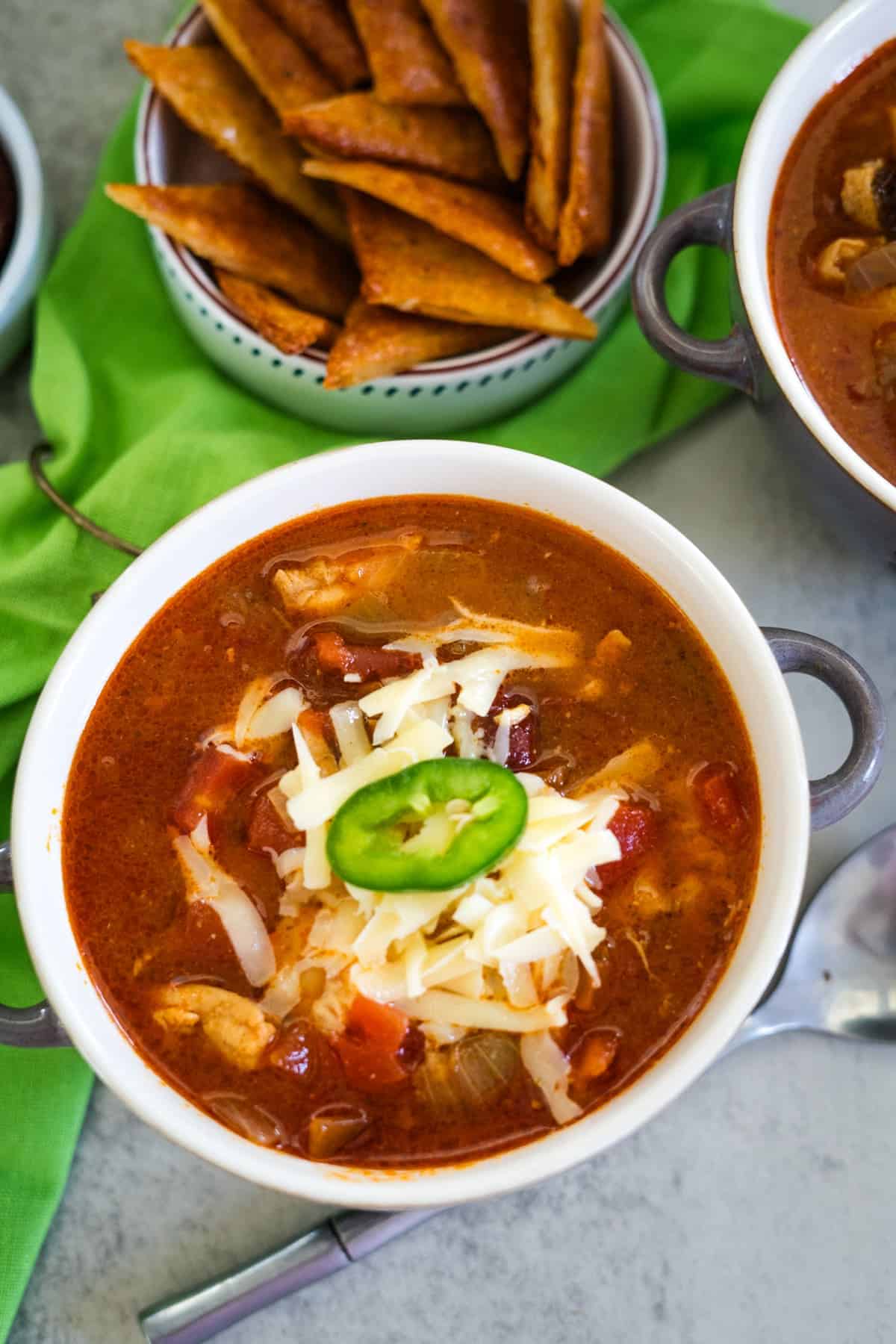 A bowl of chili topped with grated cheese and a jalapeño slice, accompanied by a bowl of chips on a green cloth.