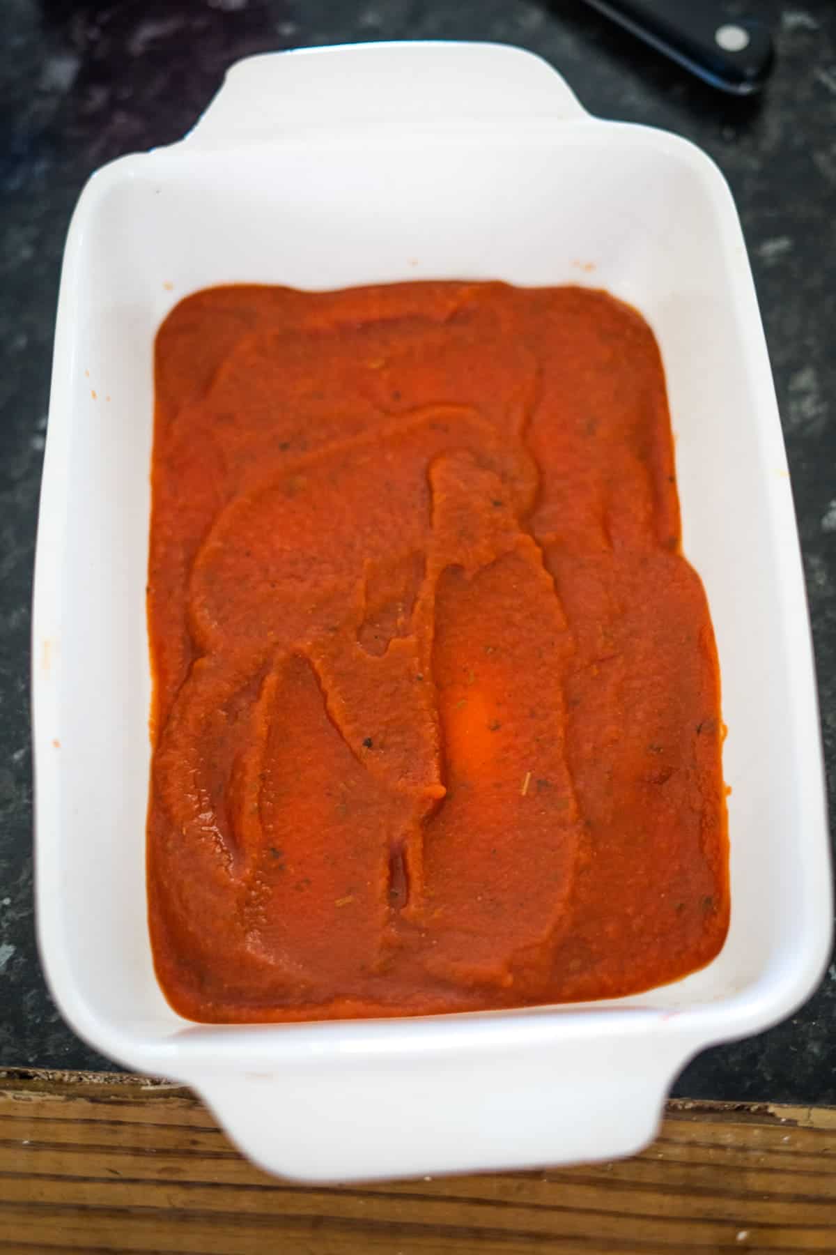 A white ceramic dish filled with rich keto spinach lasagna layered perfectly with vibrant red tomato sauce rests elegantly on a dark countertop.